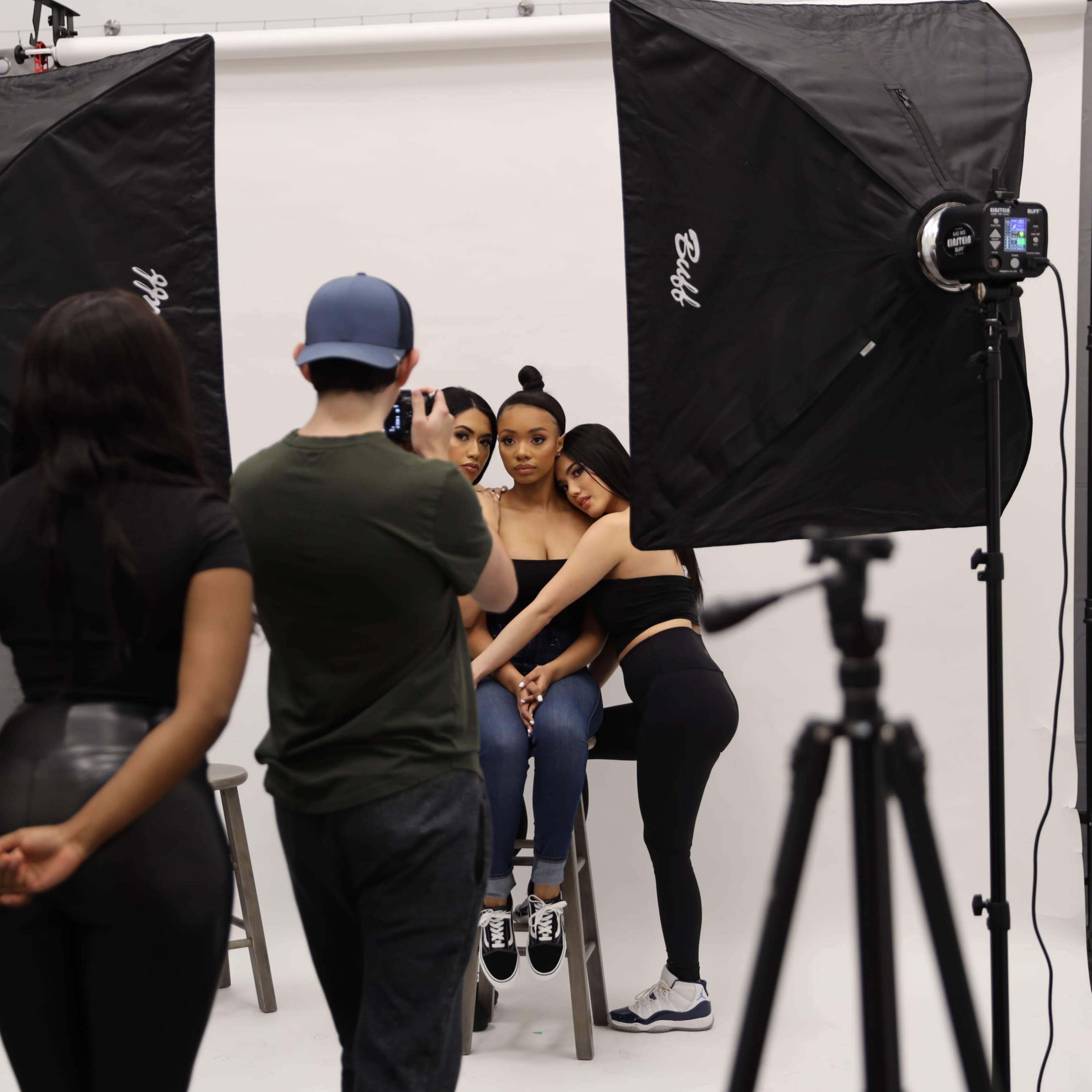 A photographer is capturing a photo of two models seated on a stool in a studio, while a third model stands behind them.