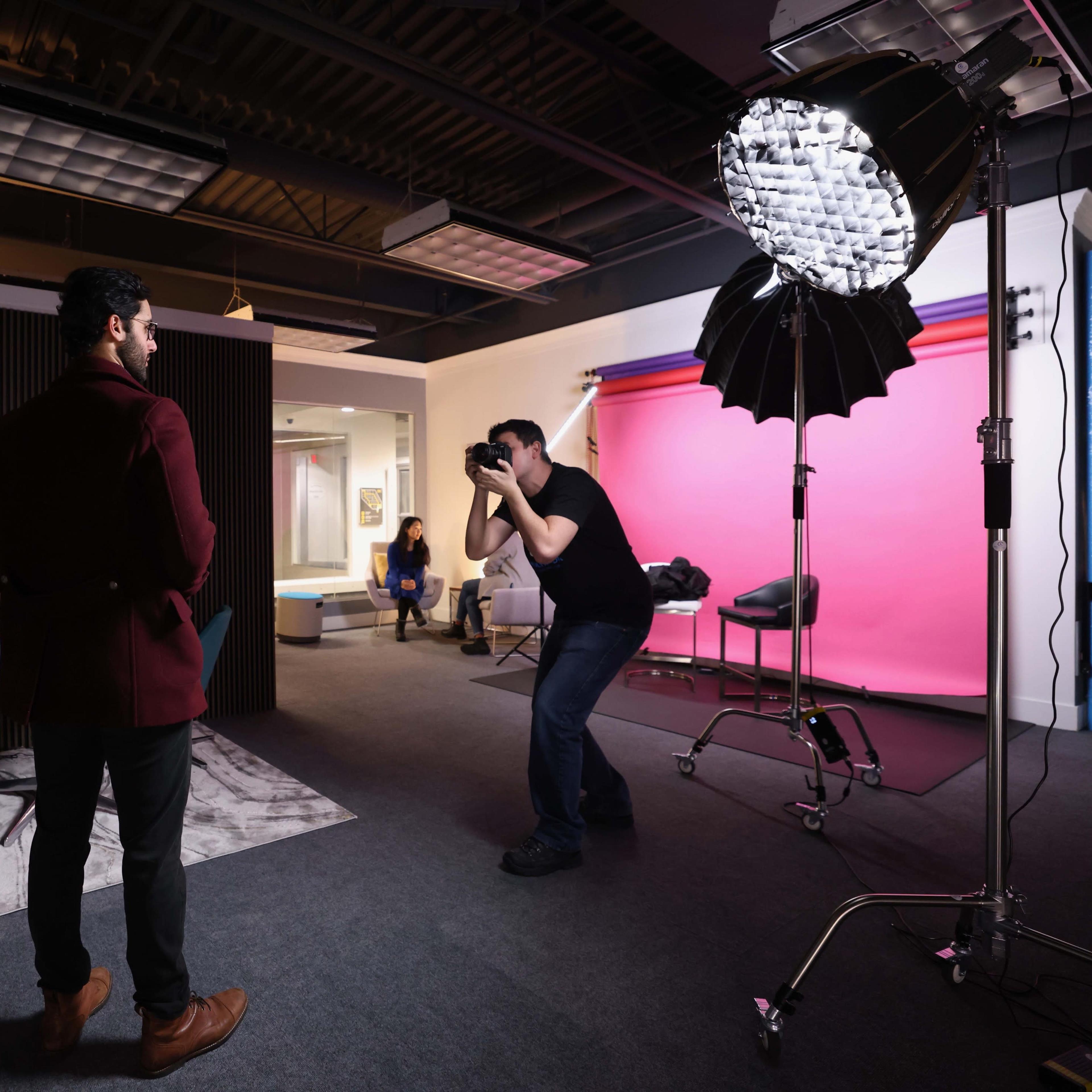 A photographer is capturing a portrait of a model in a well-lit studio filled with various backgrounds and lighting equipment.