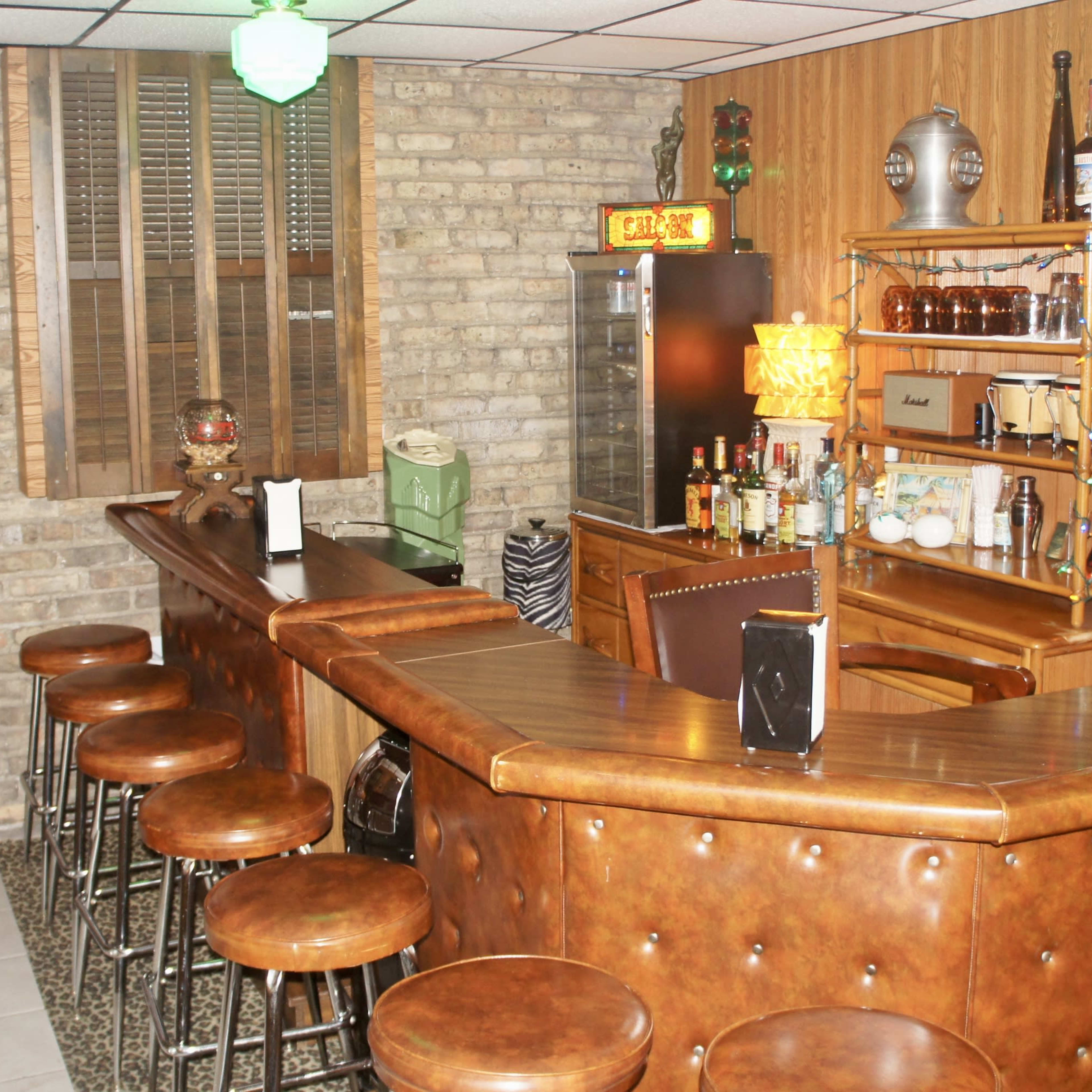 A vintage-style home bar with a curved wooden counter, leather barstools, and a brick wall backdrop.