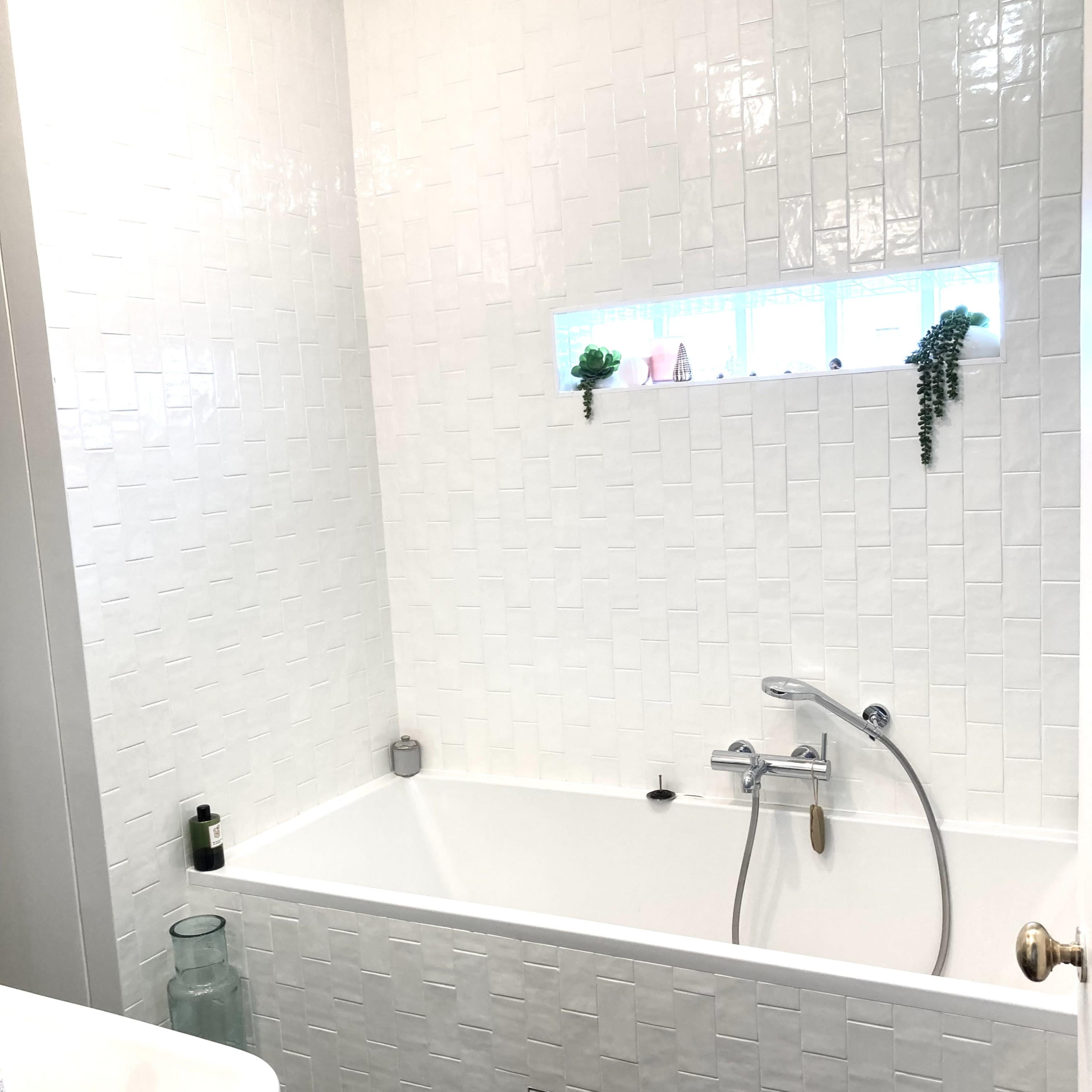 A modern bathroom featuring a white tiled wall and a bathtub with a shower fixture, complemented by a small window with decorative plants.
