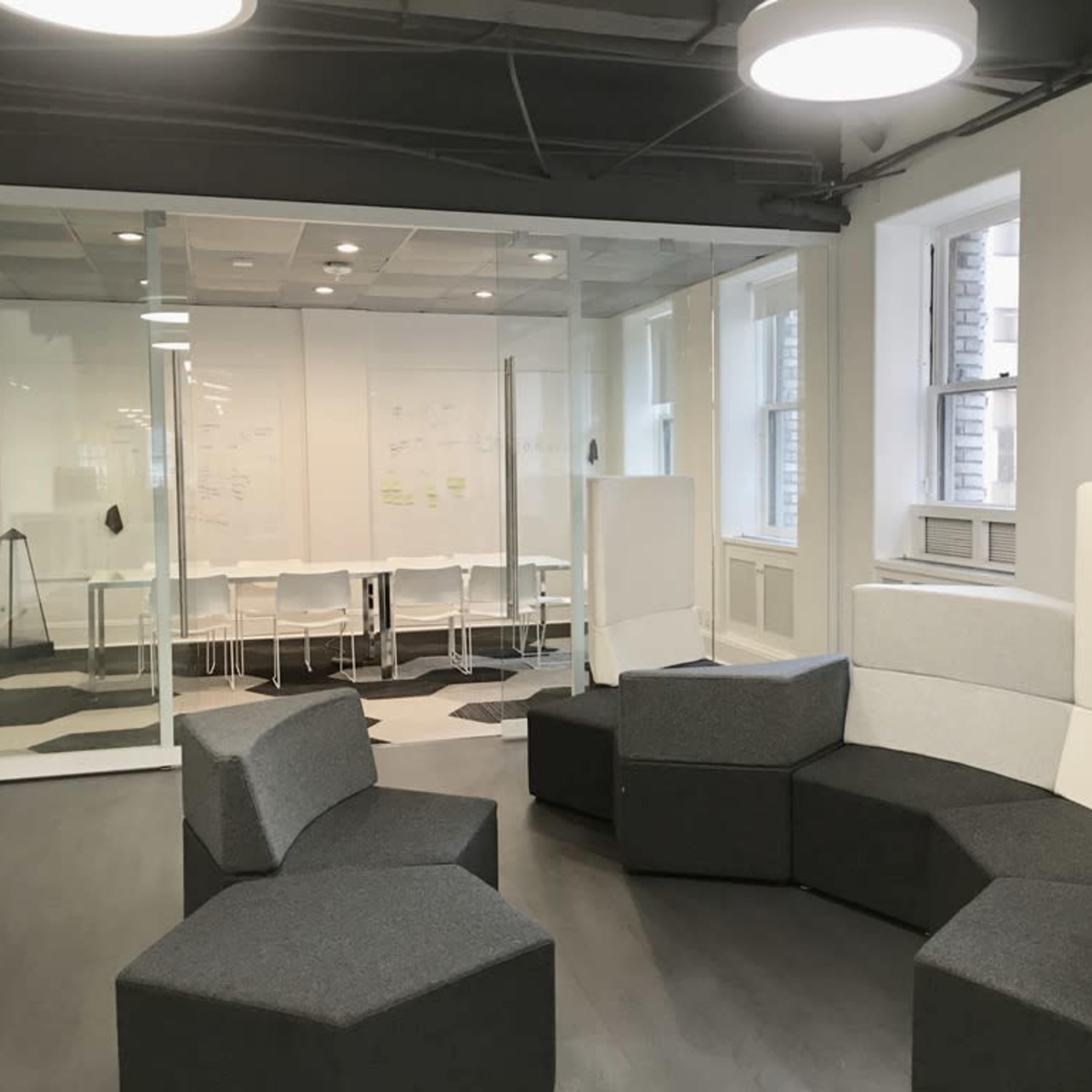 A modern office space featuring a seating area with modular gray furniture, adjacent to a glass-walled meeting room equipped with a table and chairs.