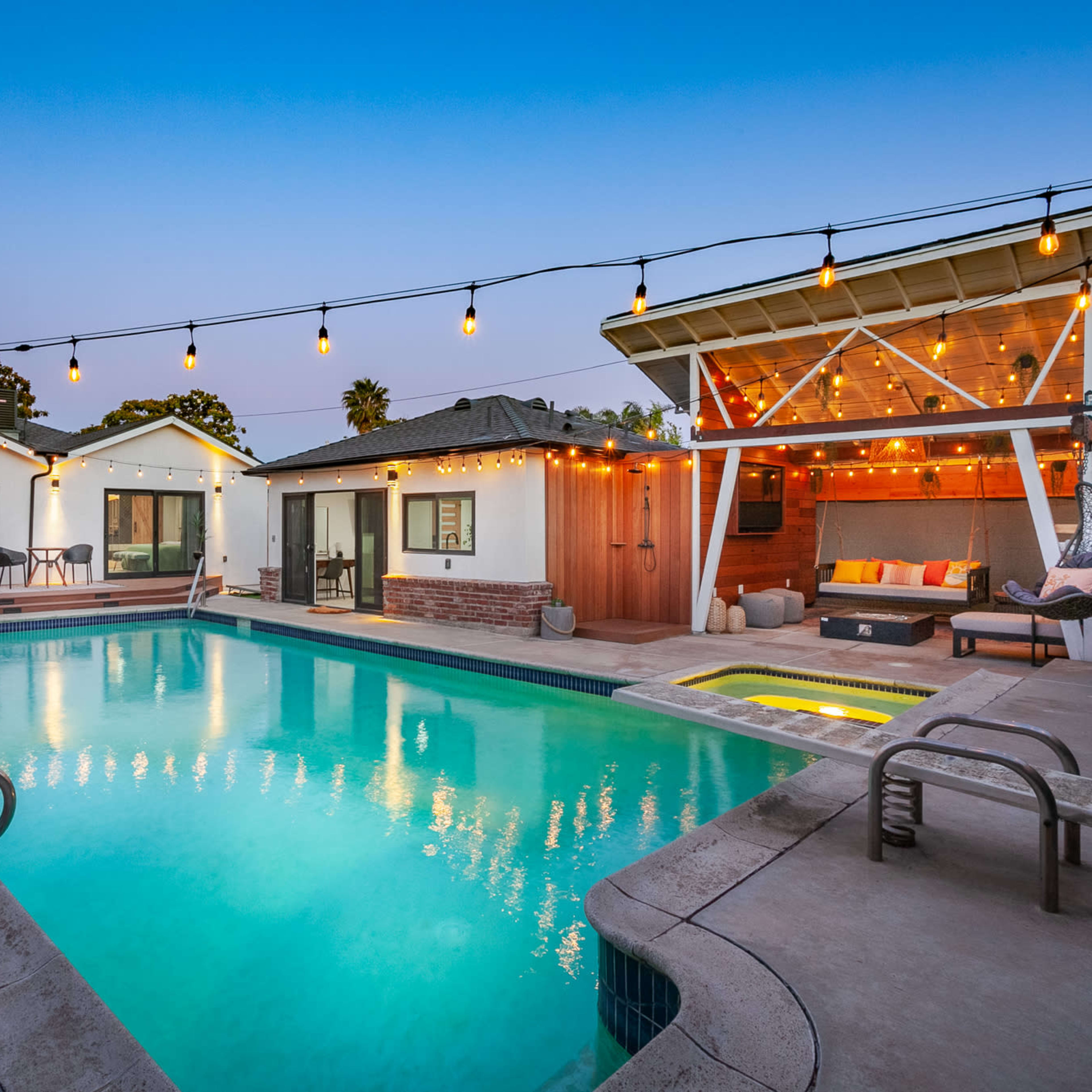 A well-lit backyard features a swimming pool surrounded by modern seating areas and a covered patio with string lights.