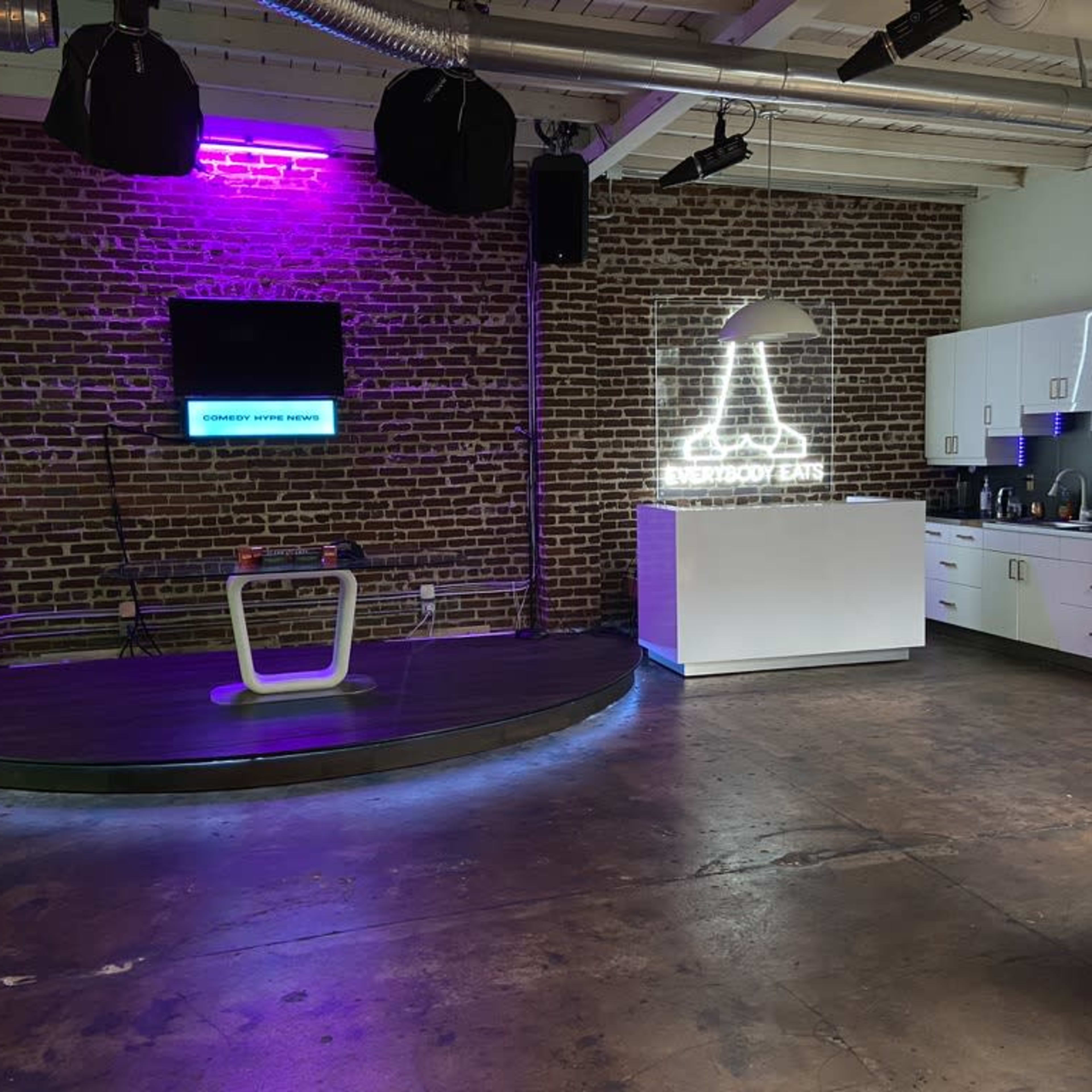 A modern interior space with exposed brick walls, featuring a circular stage area, a kitchen with white cabinetry, and neon lighting accents.