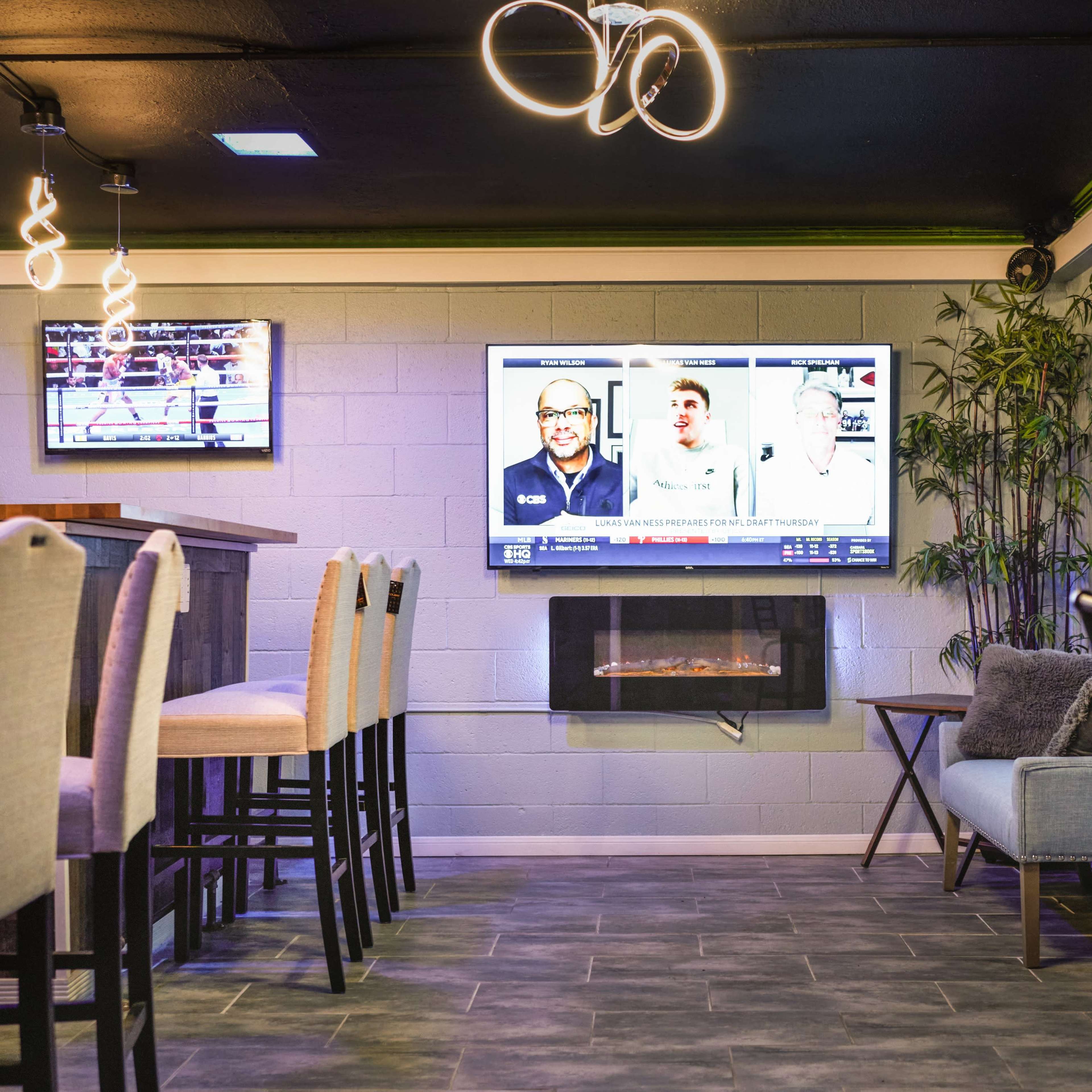 A modern bar area with a long counter, high stools, and multiple TVs displaying sports on the walls.