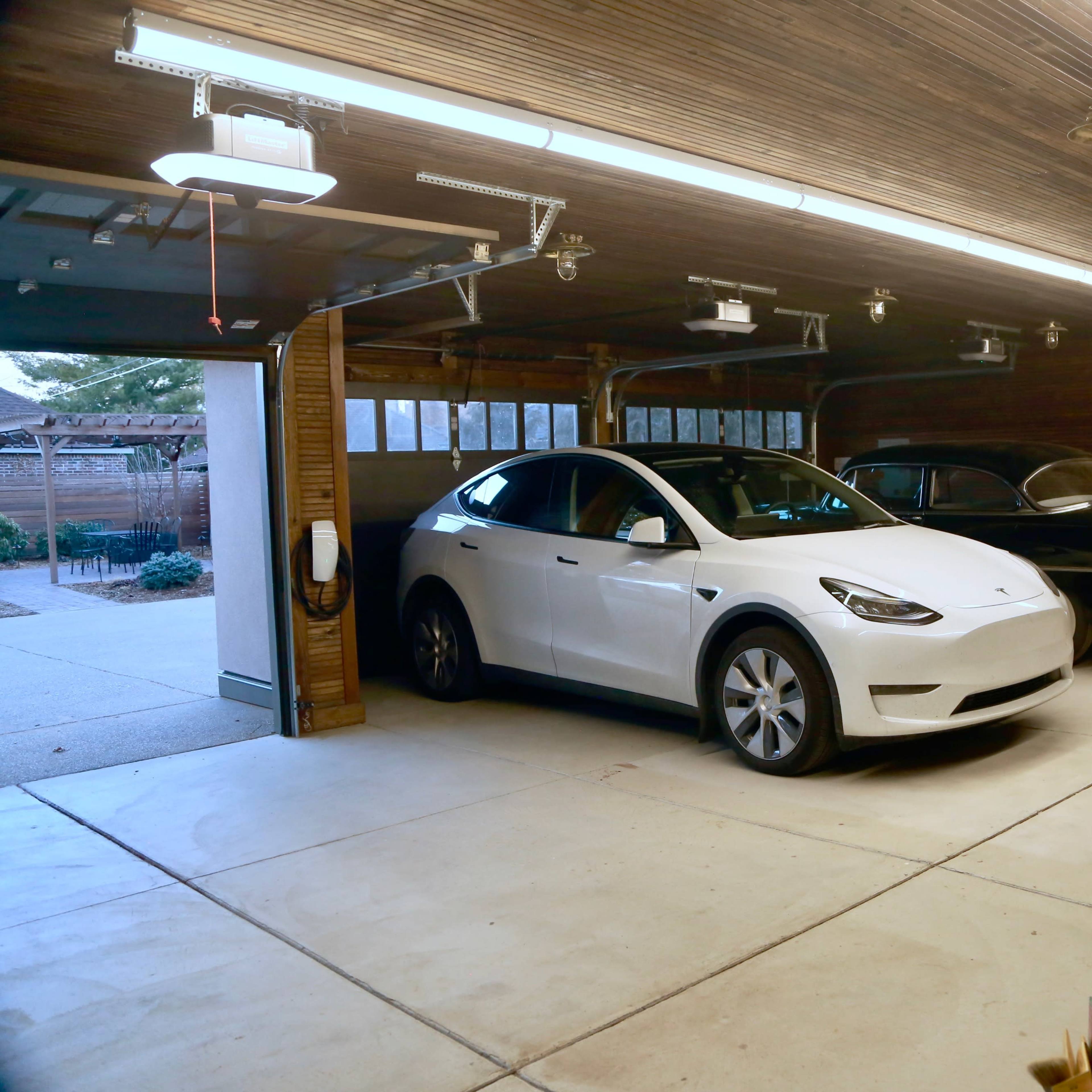 A two-car garage with a white Tesla Model Y parked next to a classic black car, featuring wooden walls and bright overhead lighting.