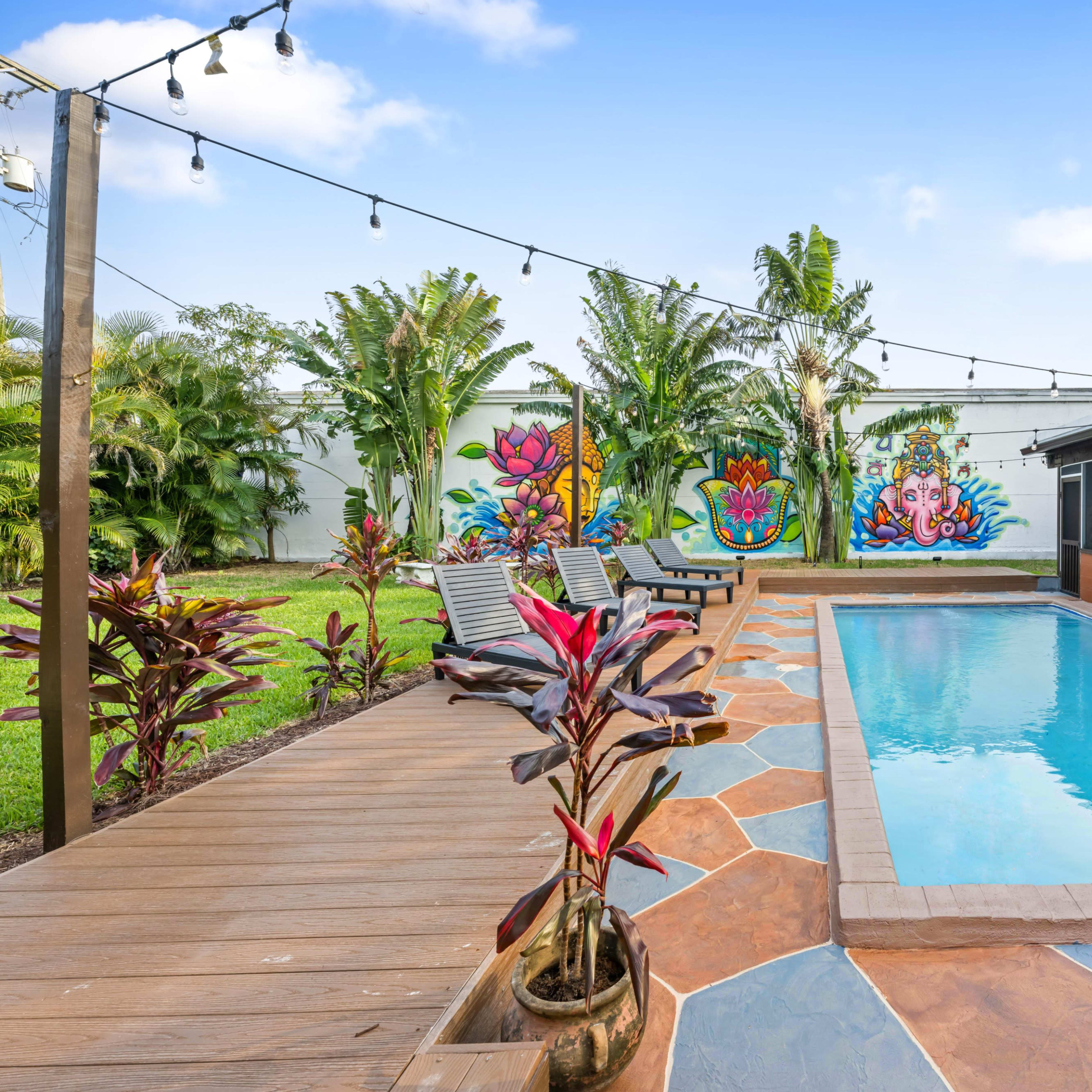 A swimming pool surrounded by a wooden deck and lounge chairs, with tropical plants and colorful murals on the wall in the background.