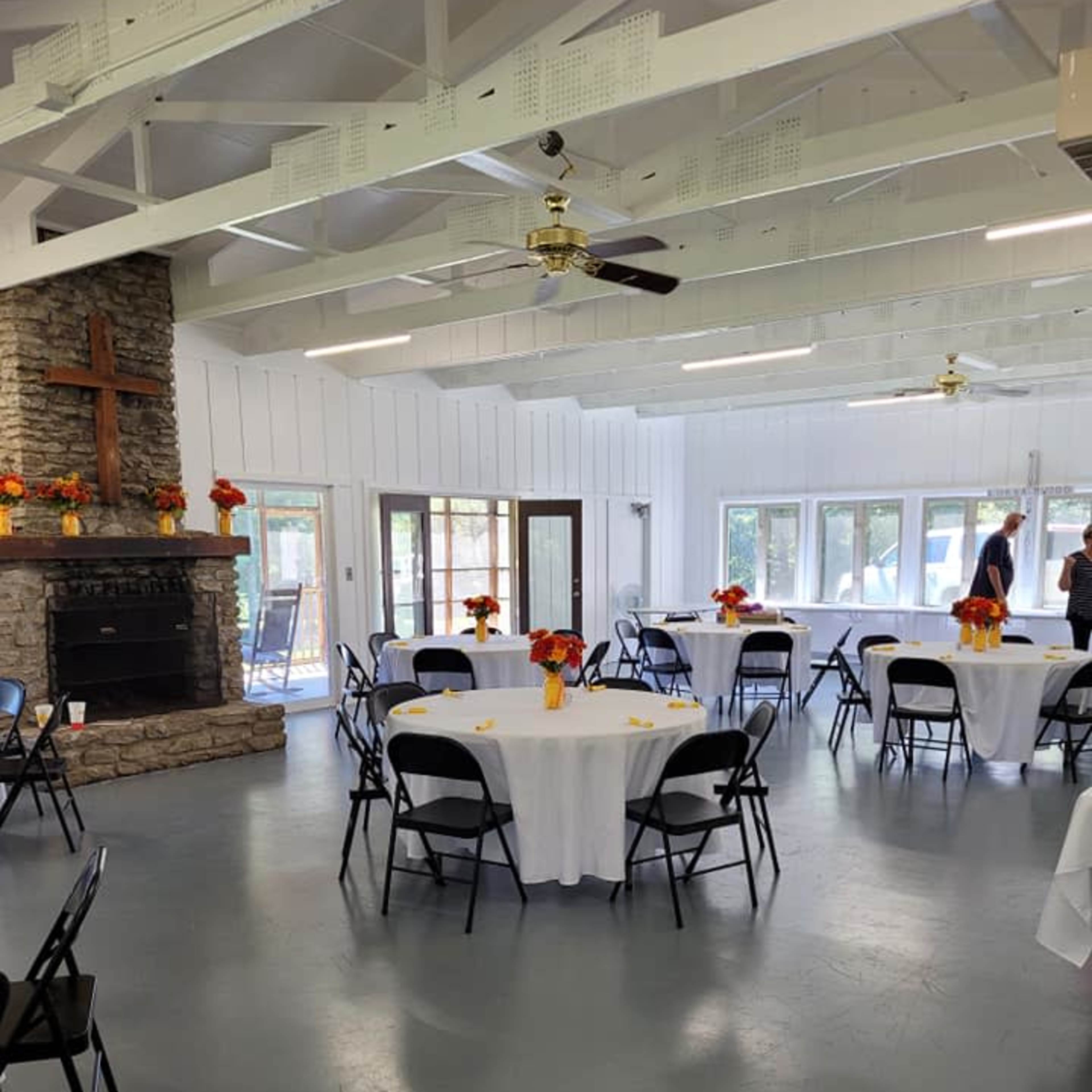 A spacious indoor venue with white walls, a stone fireplace featuring a cross, round tables covered with white tablecloths, and chairs arranged around them, all decorated with floral centerpieces.
