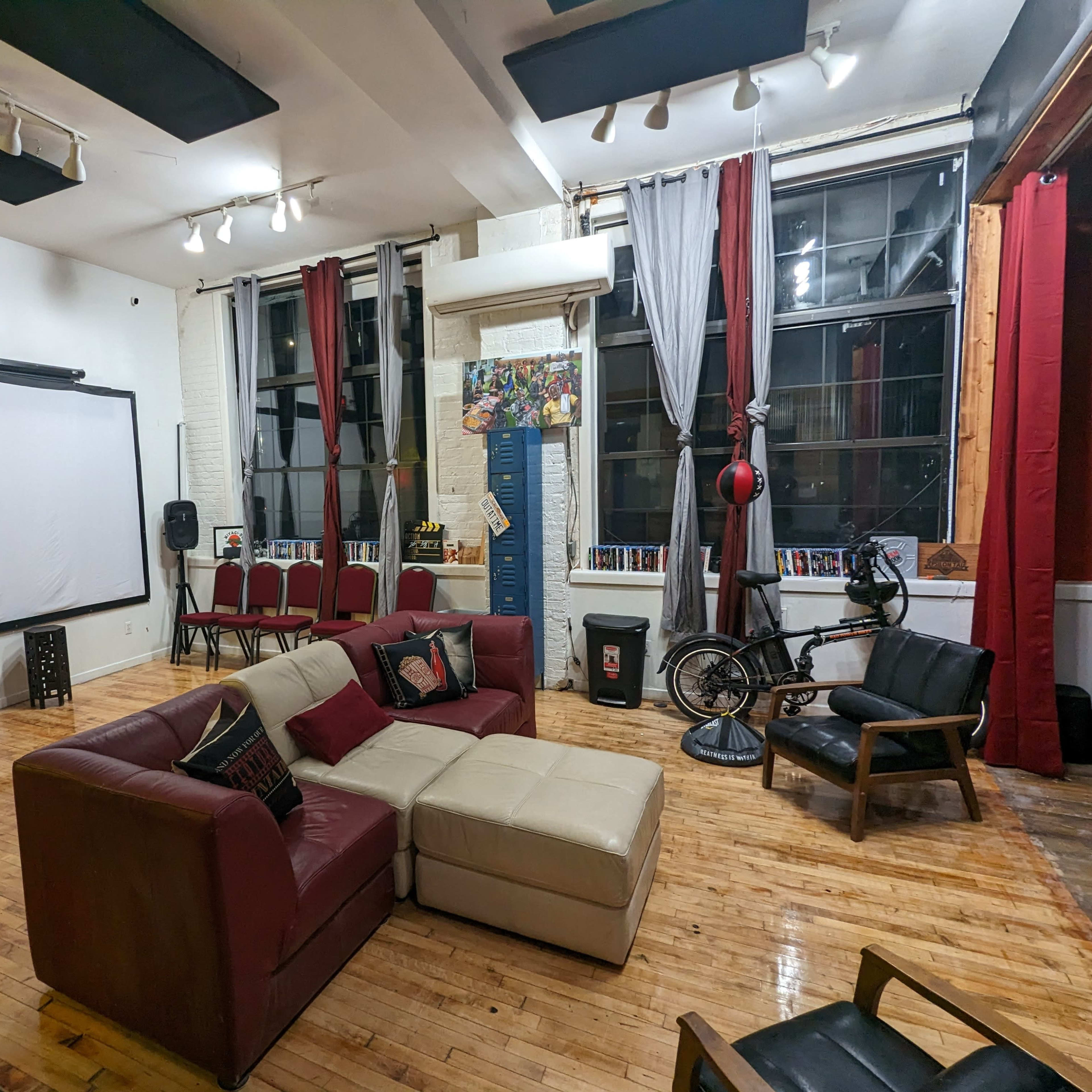 A spacious room with a large screen on one wall, seating arranged around a central couch, and various decorative elements including a bicycle and colorful curtains.