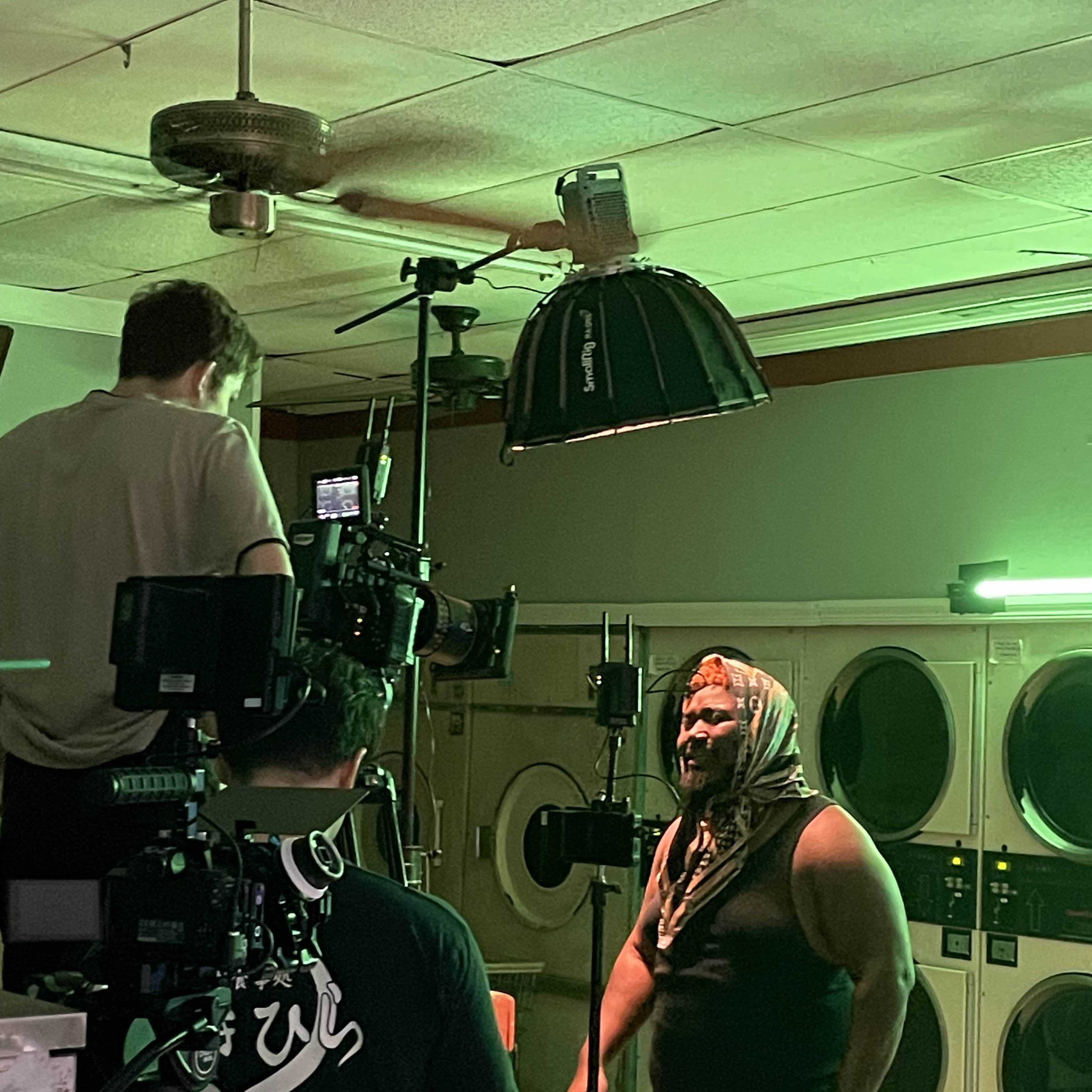 A crew films a scene featuring a man in a headscarf inside a laundry facility, with cameras and lights set up around him.