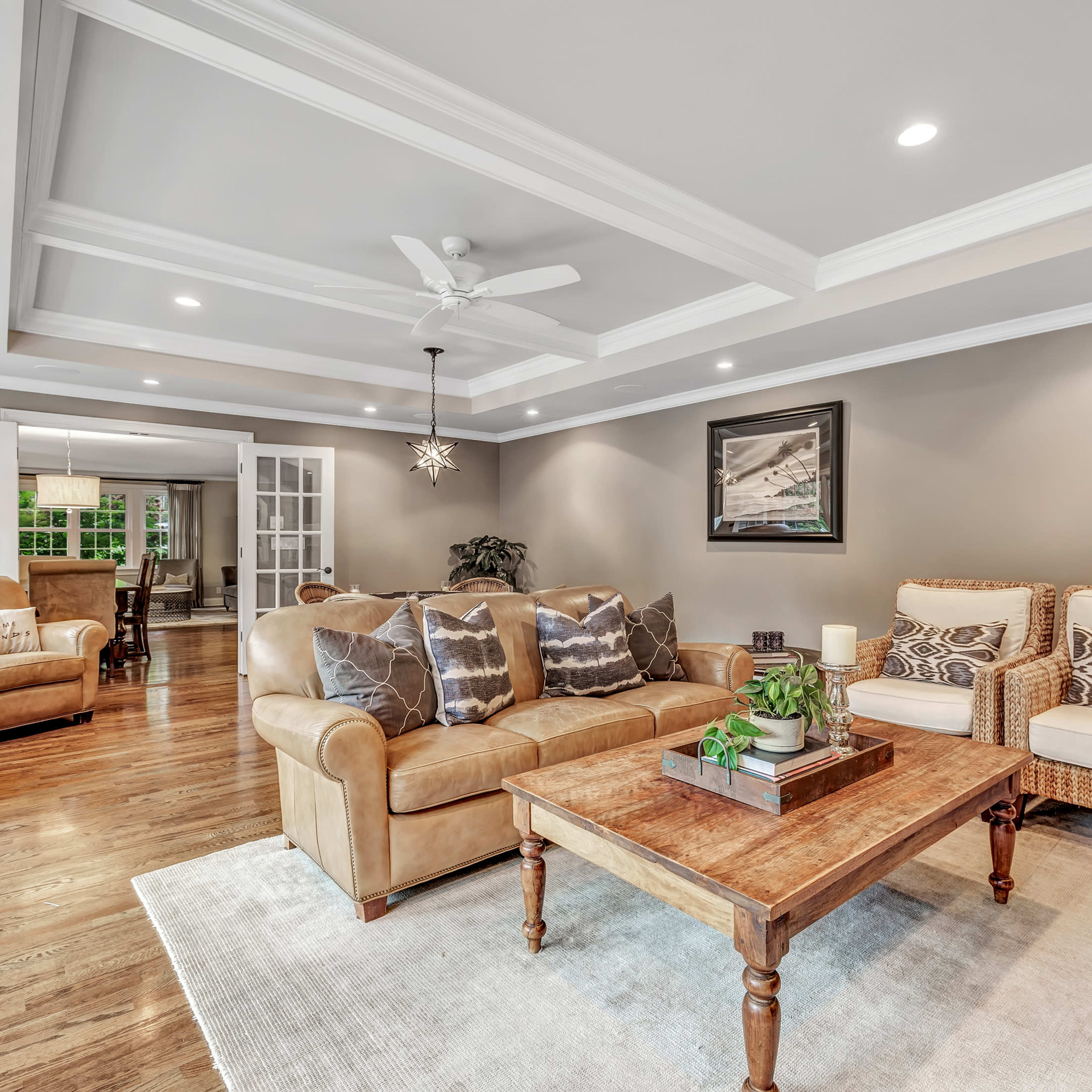 A well-lit living room features a beige couch, two patterned armchairs, a wooden coffee table, and large windows with a view into an adjoining dining area.