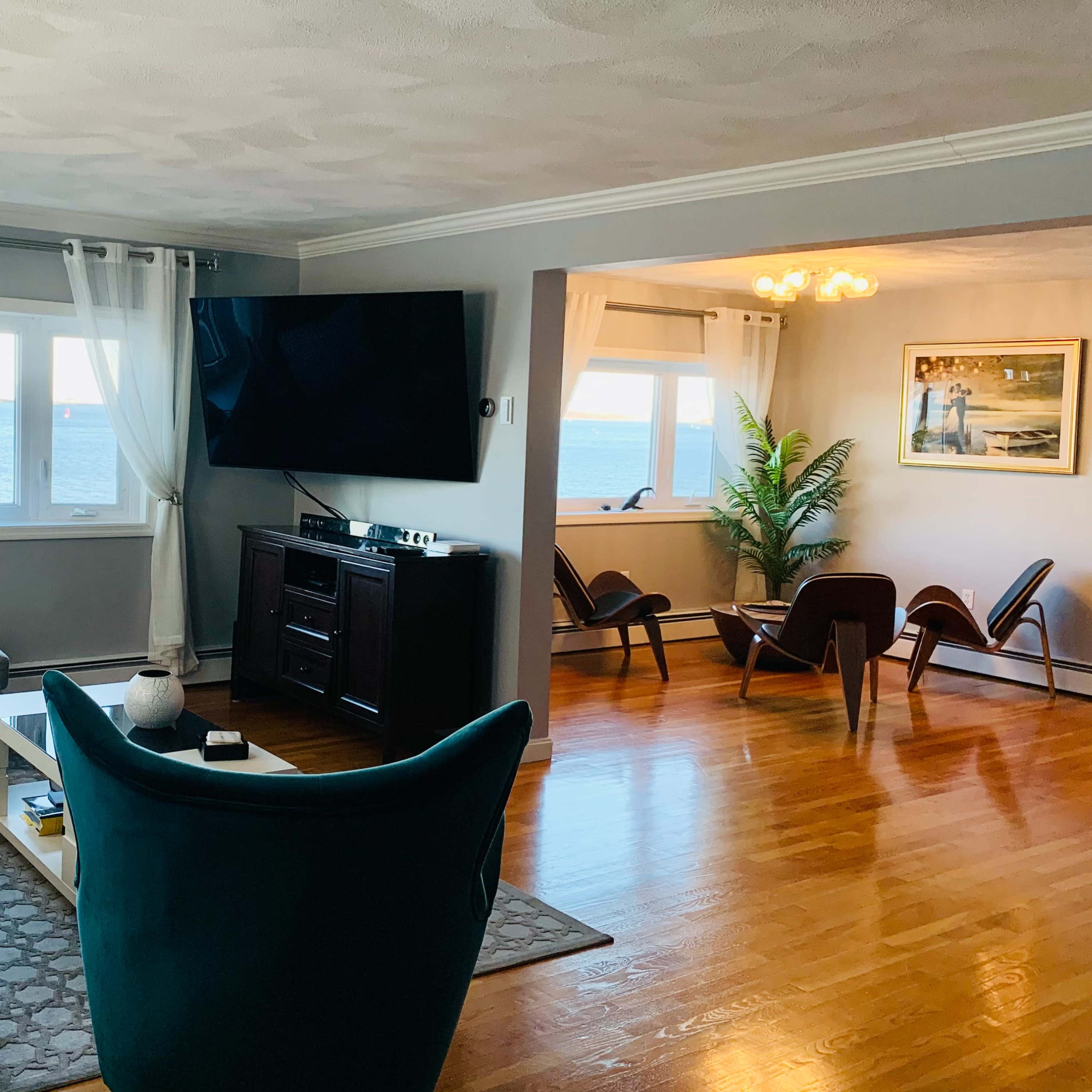 The living room features a large window overlooking the ocean, with a gray couch and a blue accent chair arranged around a coffee table and entertainment center.