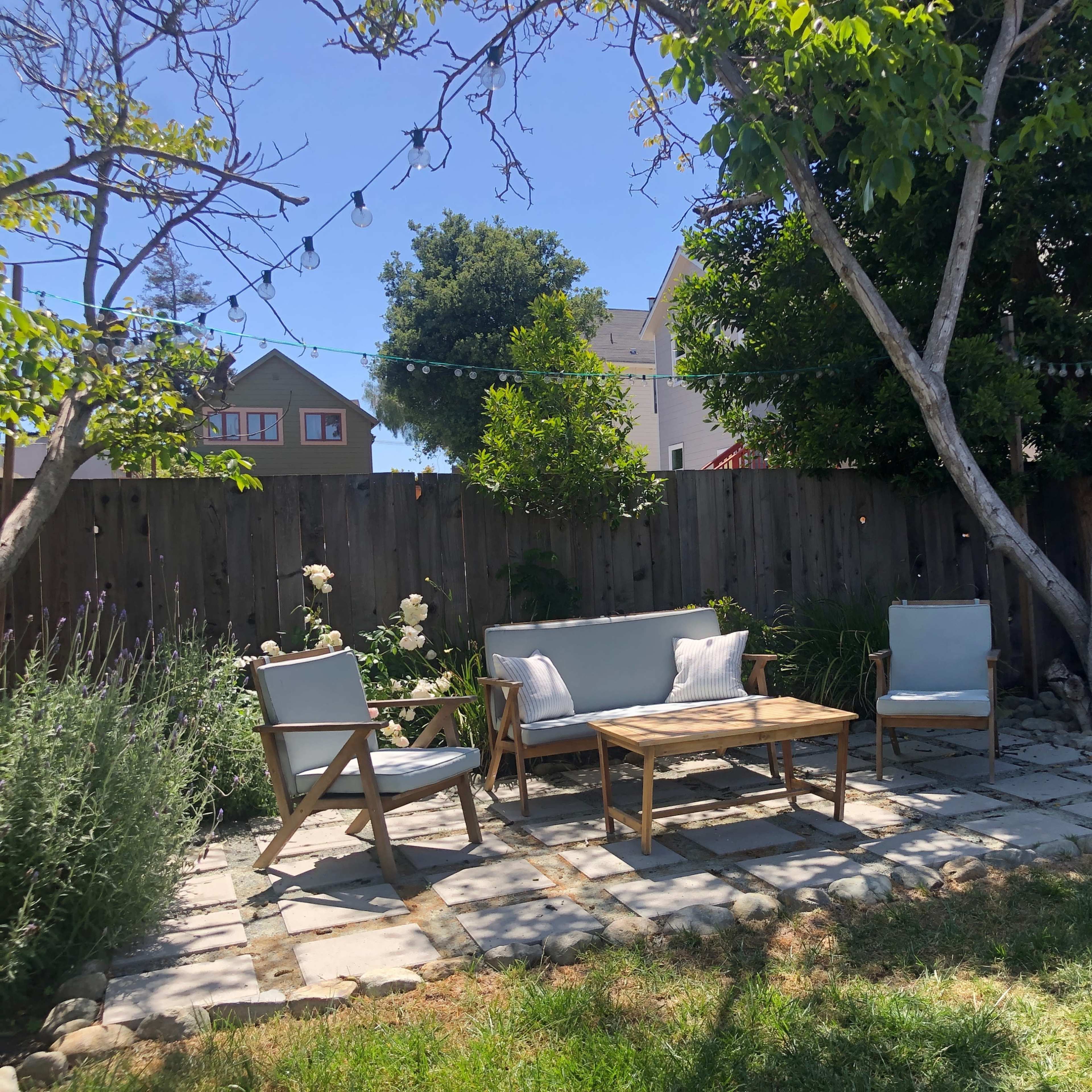 A serene backyard with a stone patio set up with a wooden table and chairs surrounded by trees and shrubs.