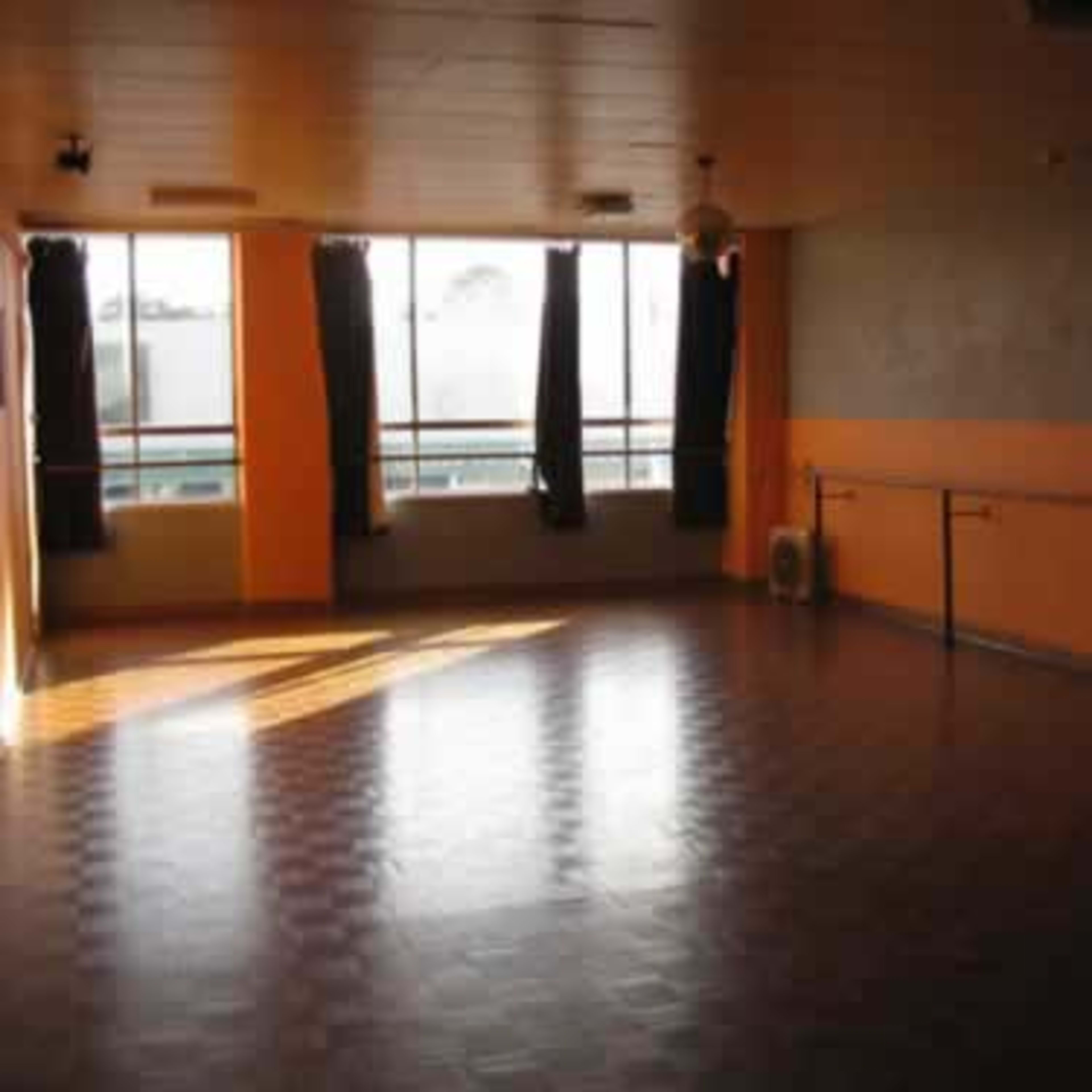 A well-lit dance studio features large windows, orange walls, and a mirrored wall along with a wooden floor and ballet barre.