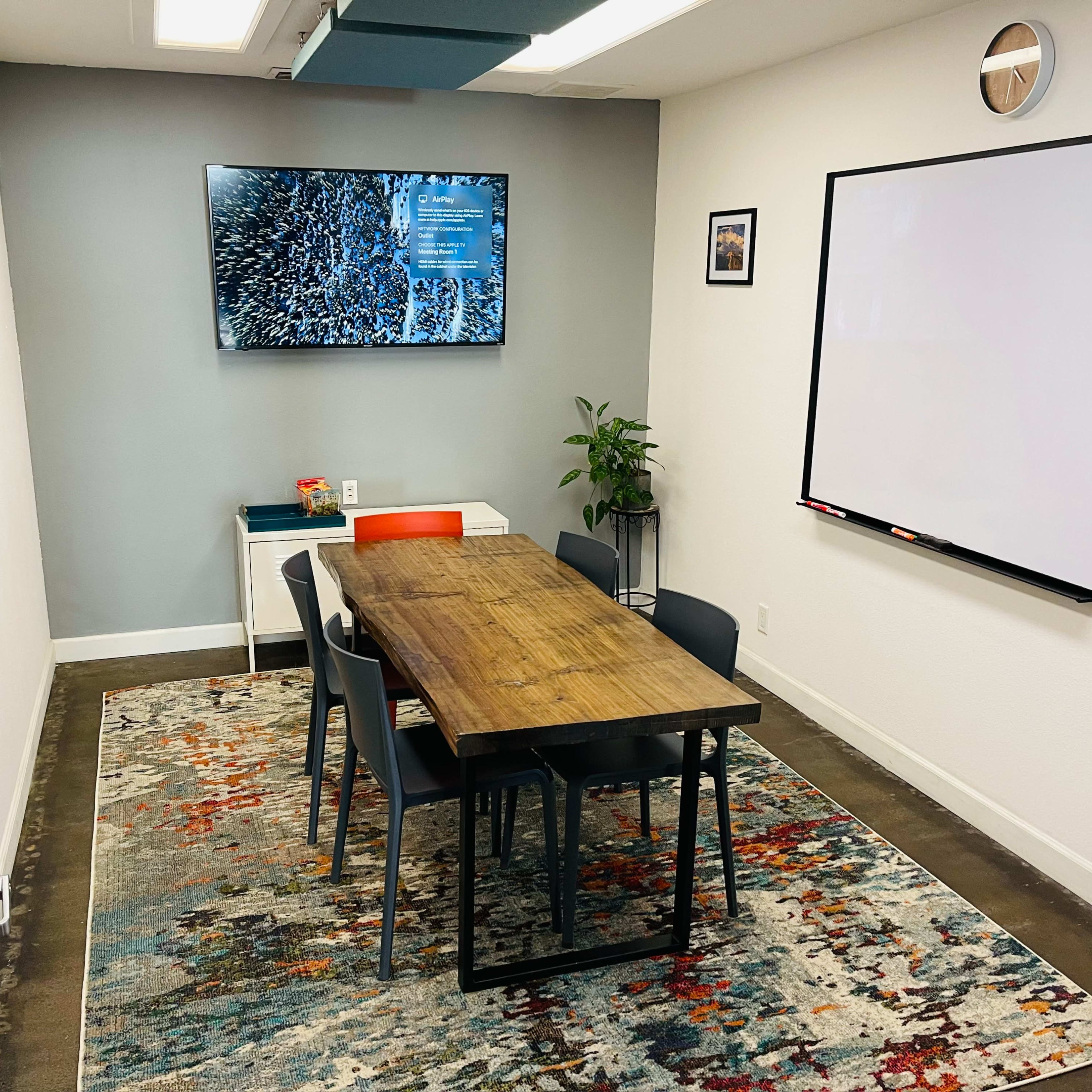 A small conference room features a wooden table surrounded by chairs, a large TV on the wall, a whiteboard, and a plant in the corner.