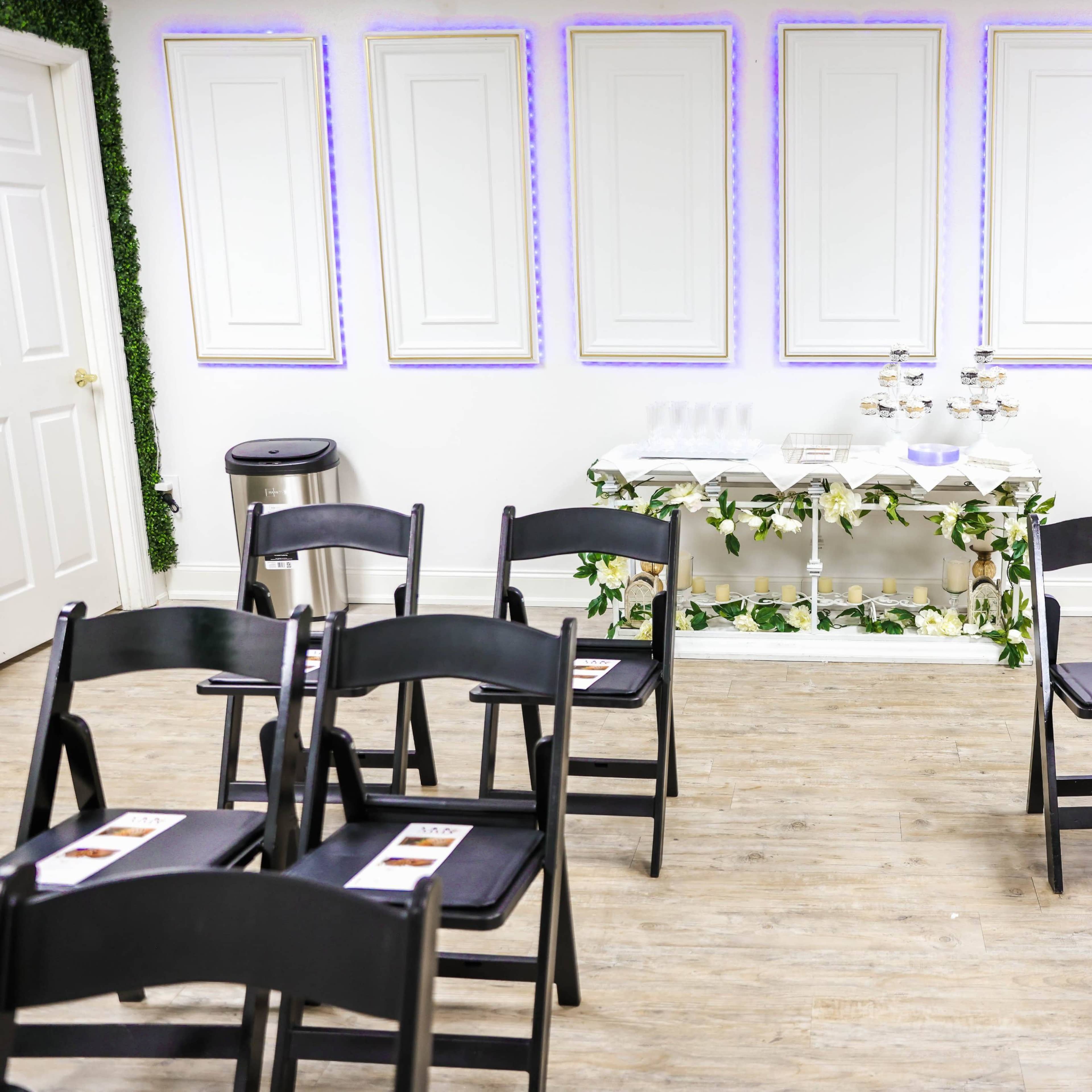 A rows of black folding chairs face a decorative table with refreshments in a room featuring white walls and framed panels.