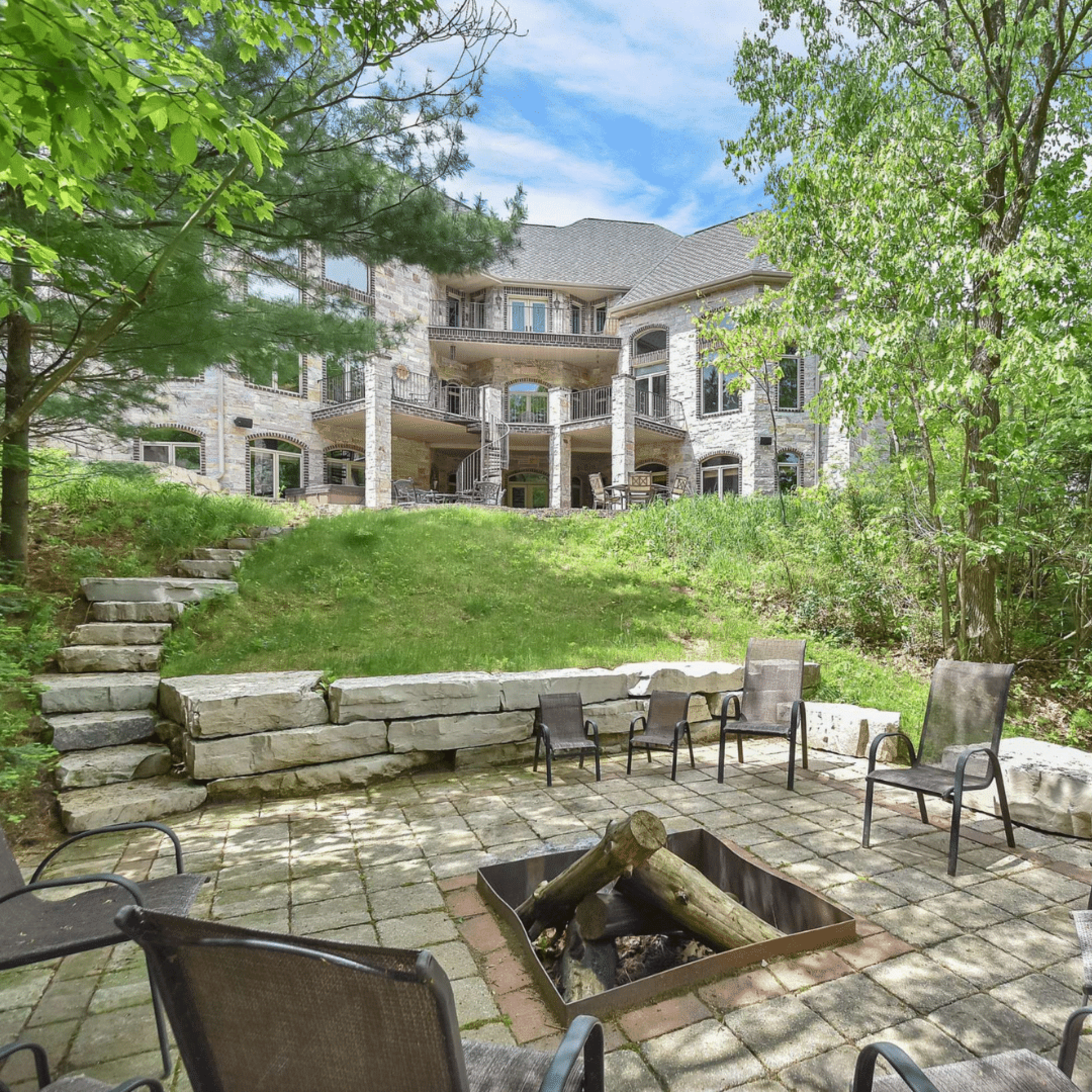 A stone fire pit surrounded by chairs, with a spacious house and green trees in the background.