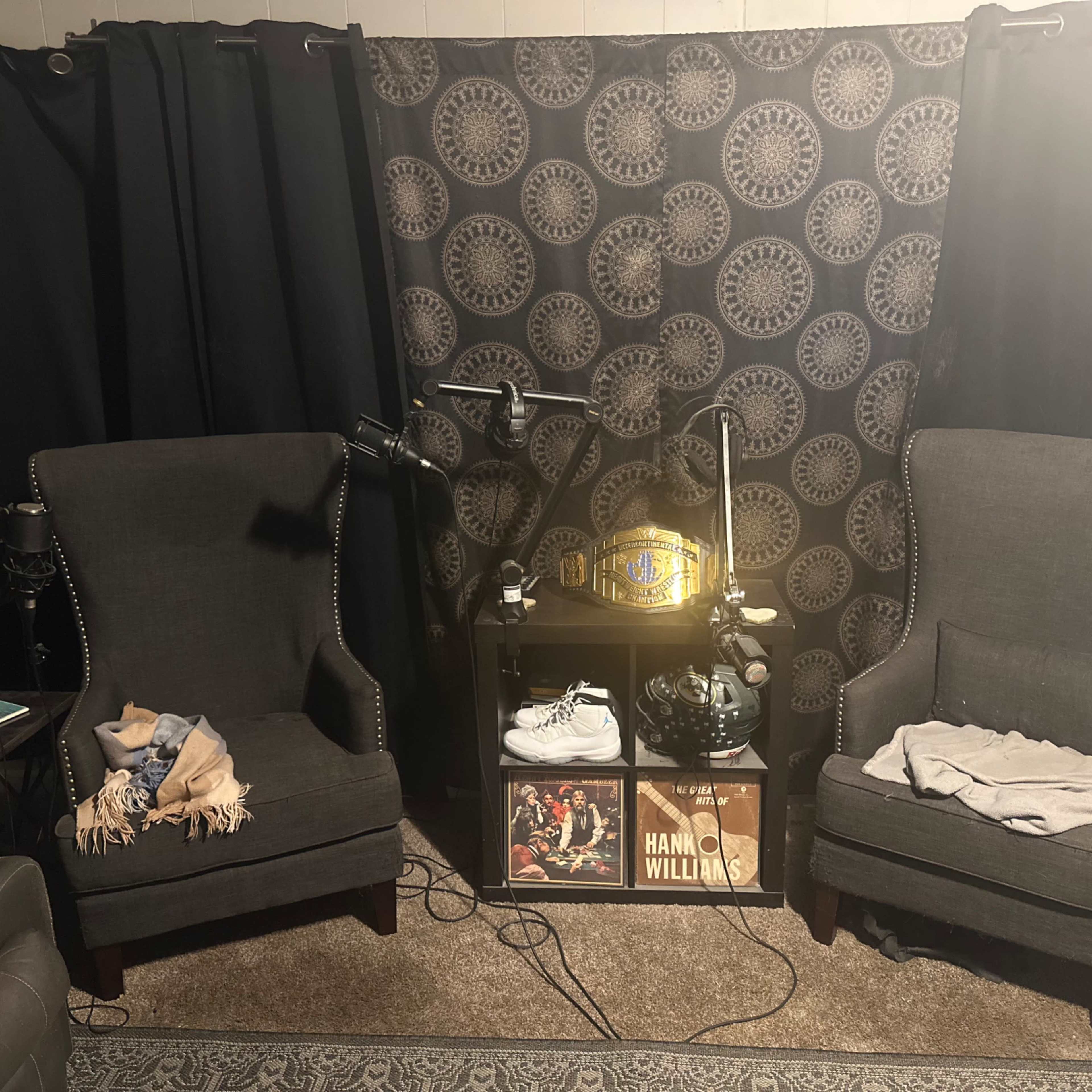 Two dark armchairs facing a small table adorned with a championship belt and some framed records, set against a patterned curtain.