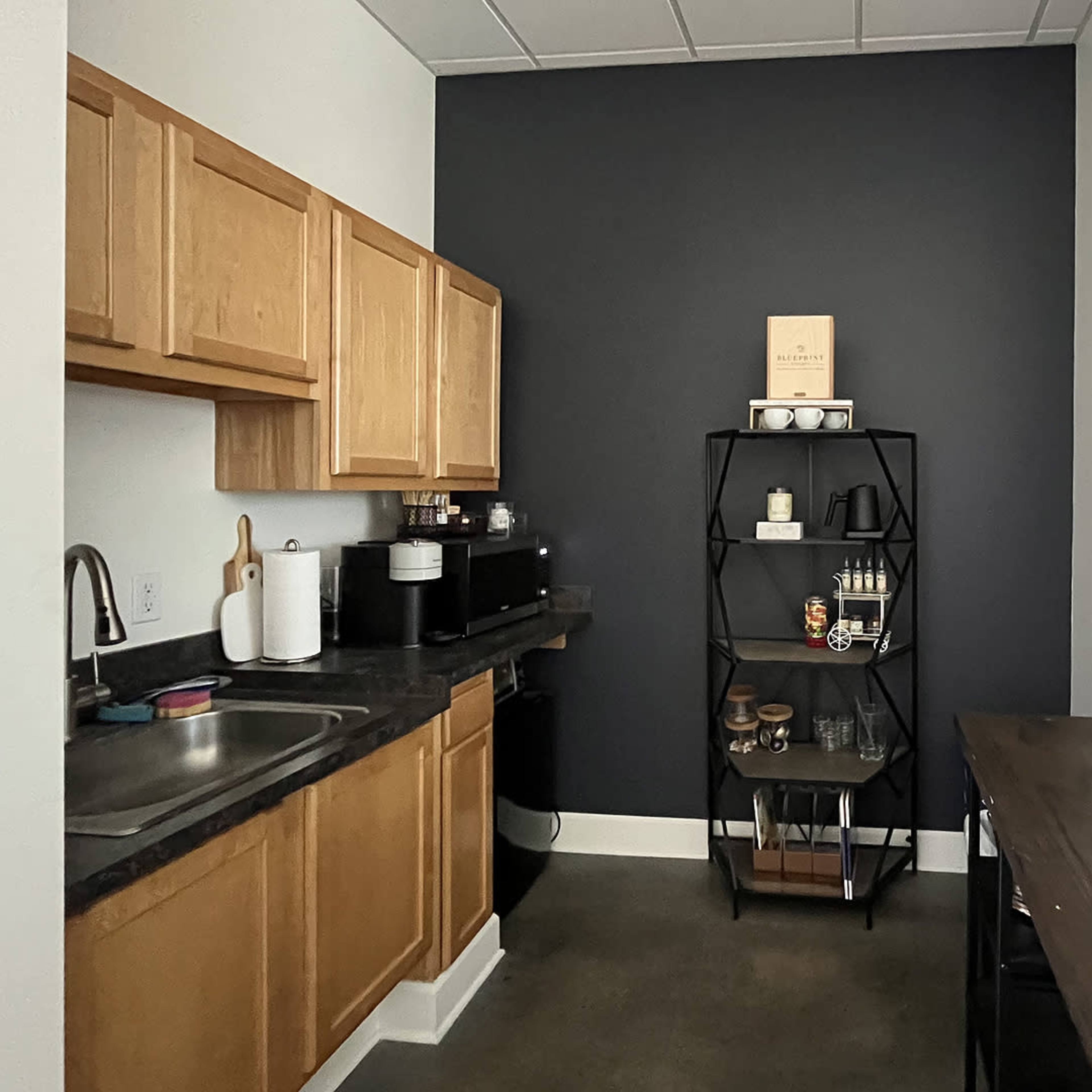 A small kitchen with wooden cabinets, a dark wall, a sink, a coffee maker, and a shelving unit displaying various kitchen items.