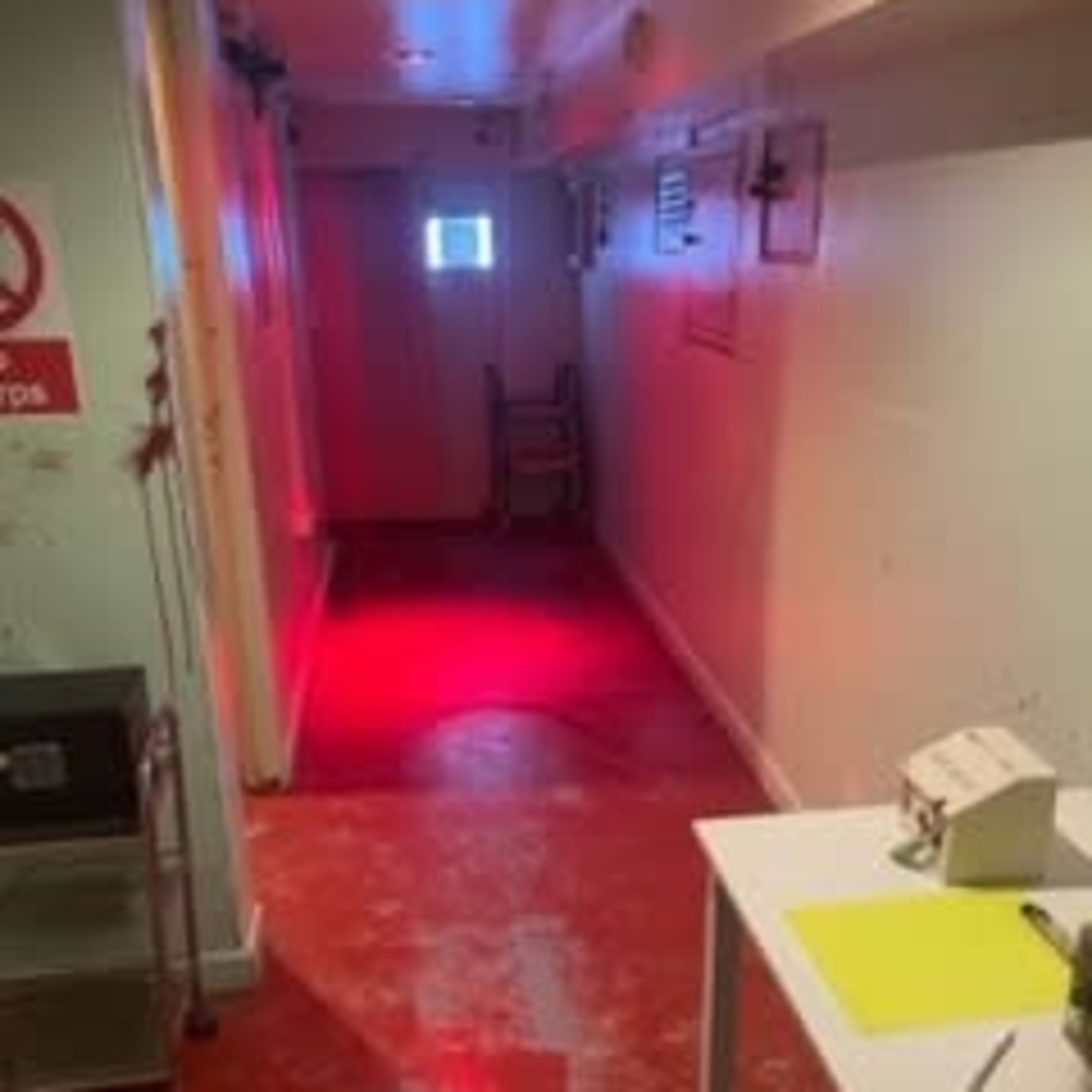 A narrow hallway with red lighting, a window at the far end, and a chair placed against the wall.