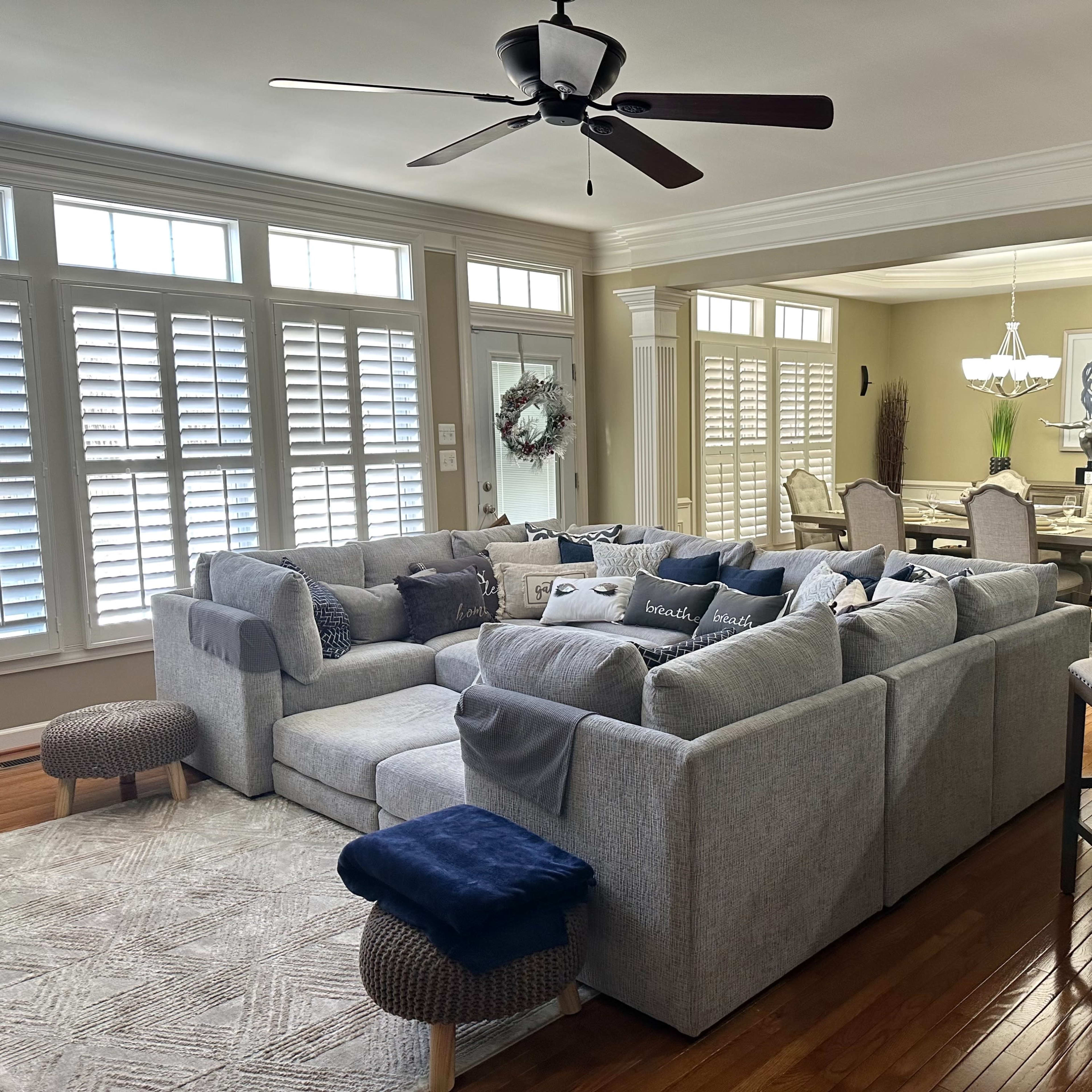 A spacious living room features a large sectional sofa with various cushions, a ceiling fan, and a view into a dining area with a chandelier and a table set for several people.