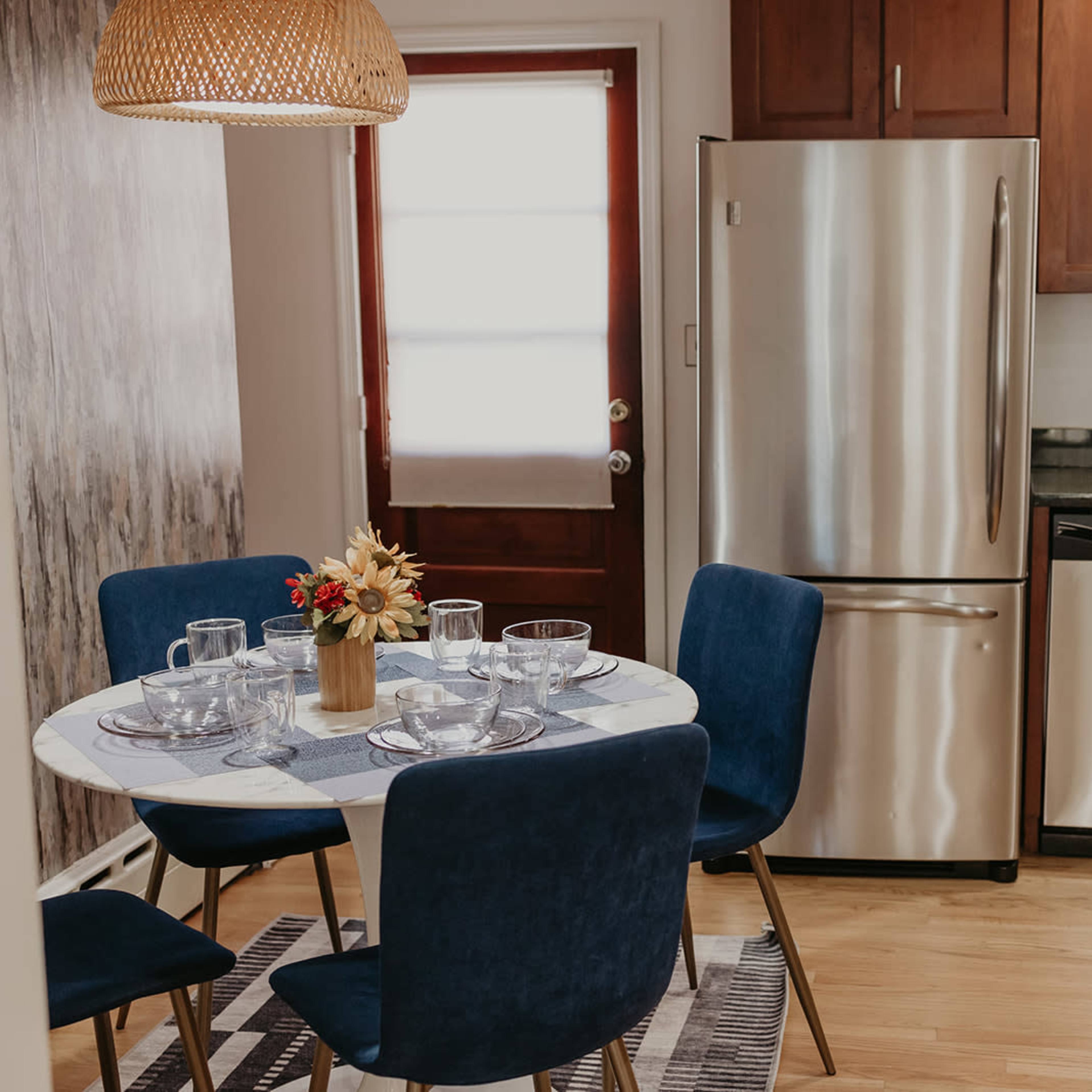A small dining area features a round table set with glassware and a floral centerpiece, surrounded by blue chairs, beside a modern kitchen with stainless steel appliances.