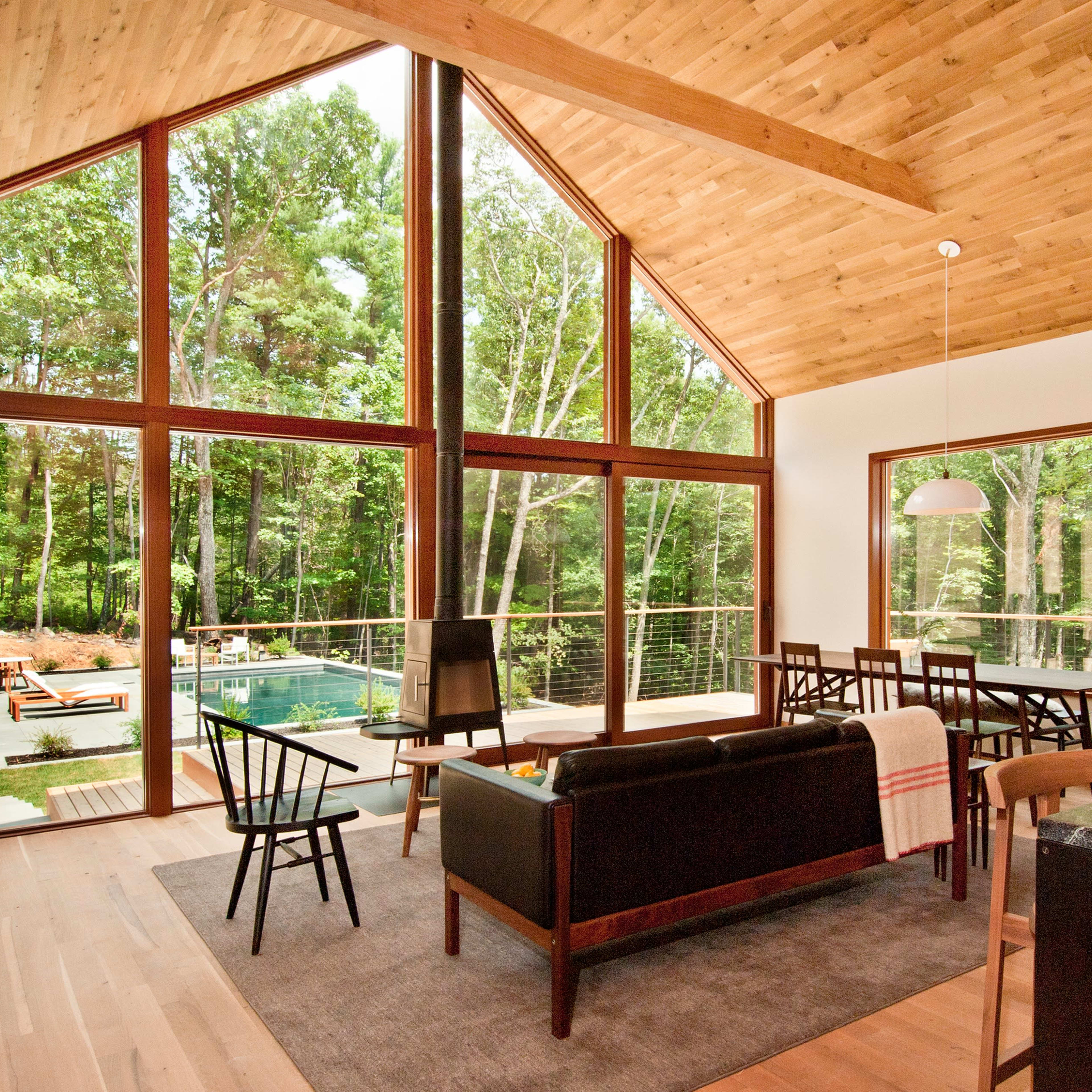 A modern living area features large windows overlooking a pool and wooded landscape, with a dining table and stylish furniture.