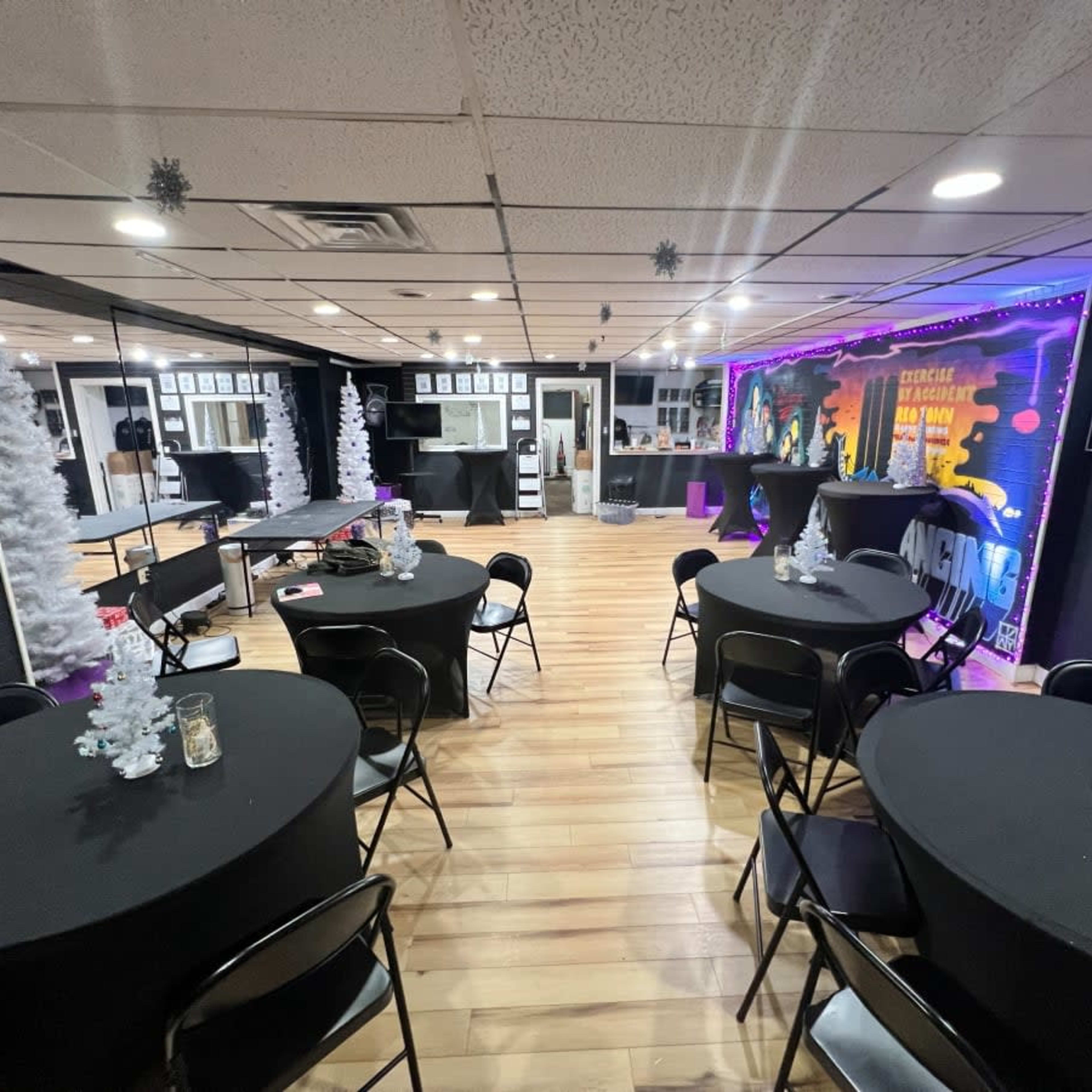 A decorated event space with black tables, small white Christmas trees, and festive lighting on the walls.