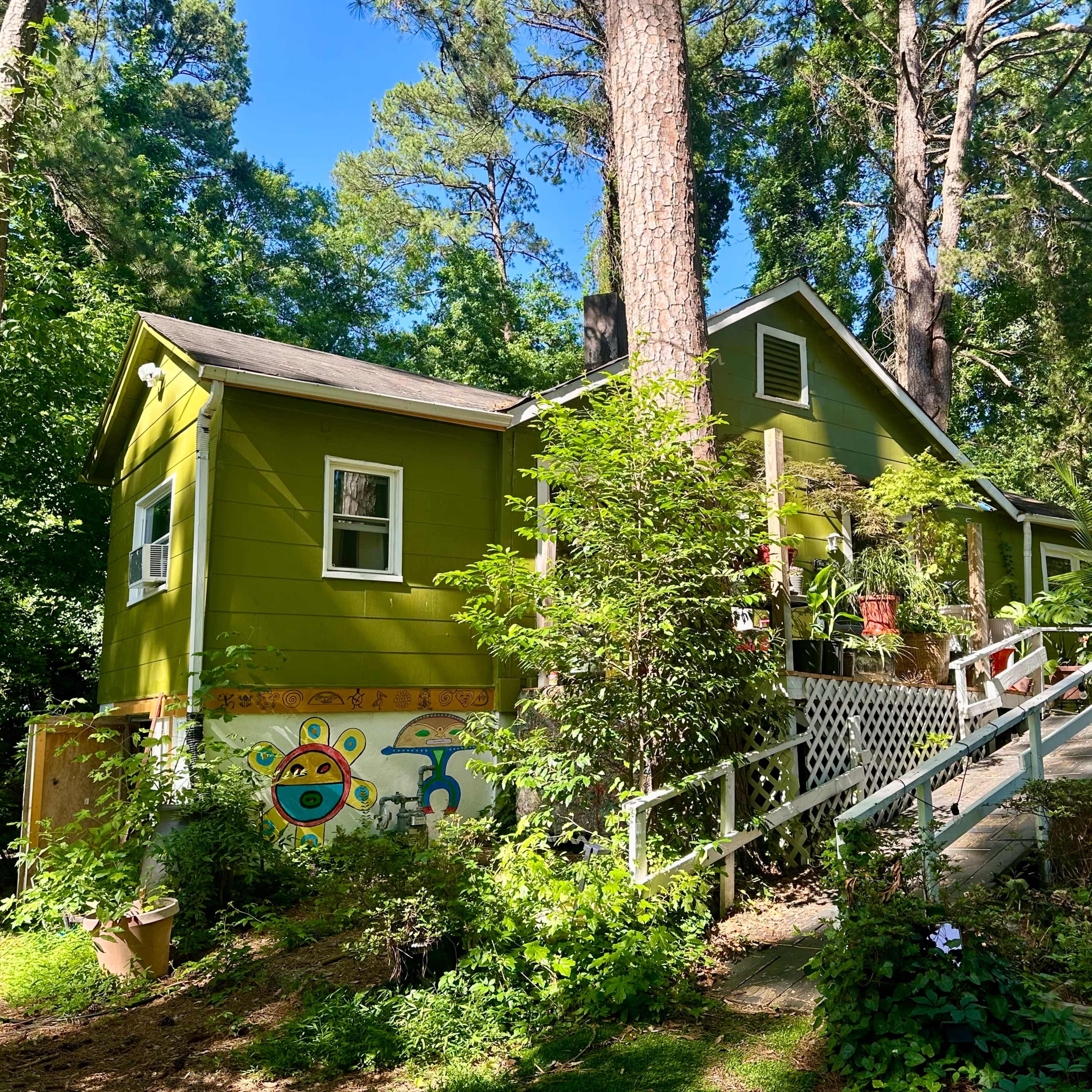 A green house with a decorative mural on one side is surrounded by trees and plants, featuring a front ramp leading to the entrance.