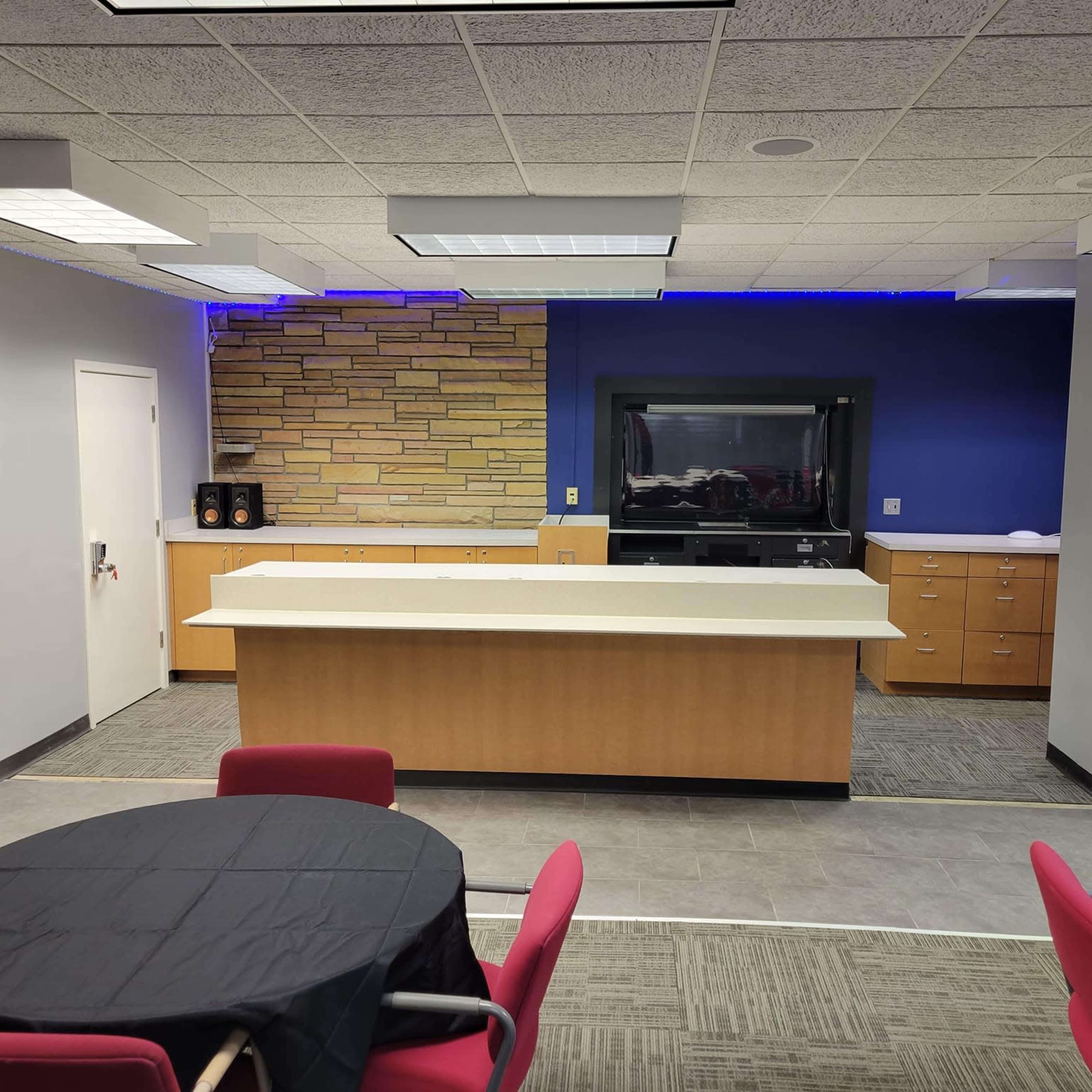 A modern kitchen area in a conference or meeting room, featuring a large countertop, cabinetry, and a television against a blue and stone wall.