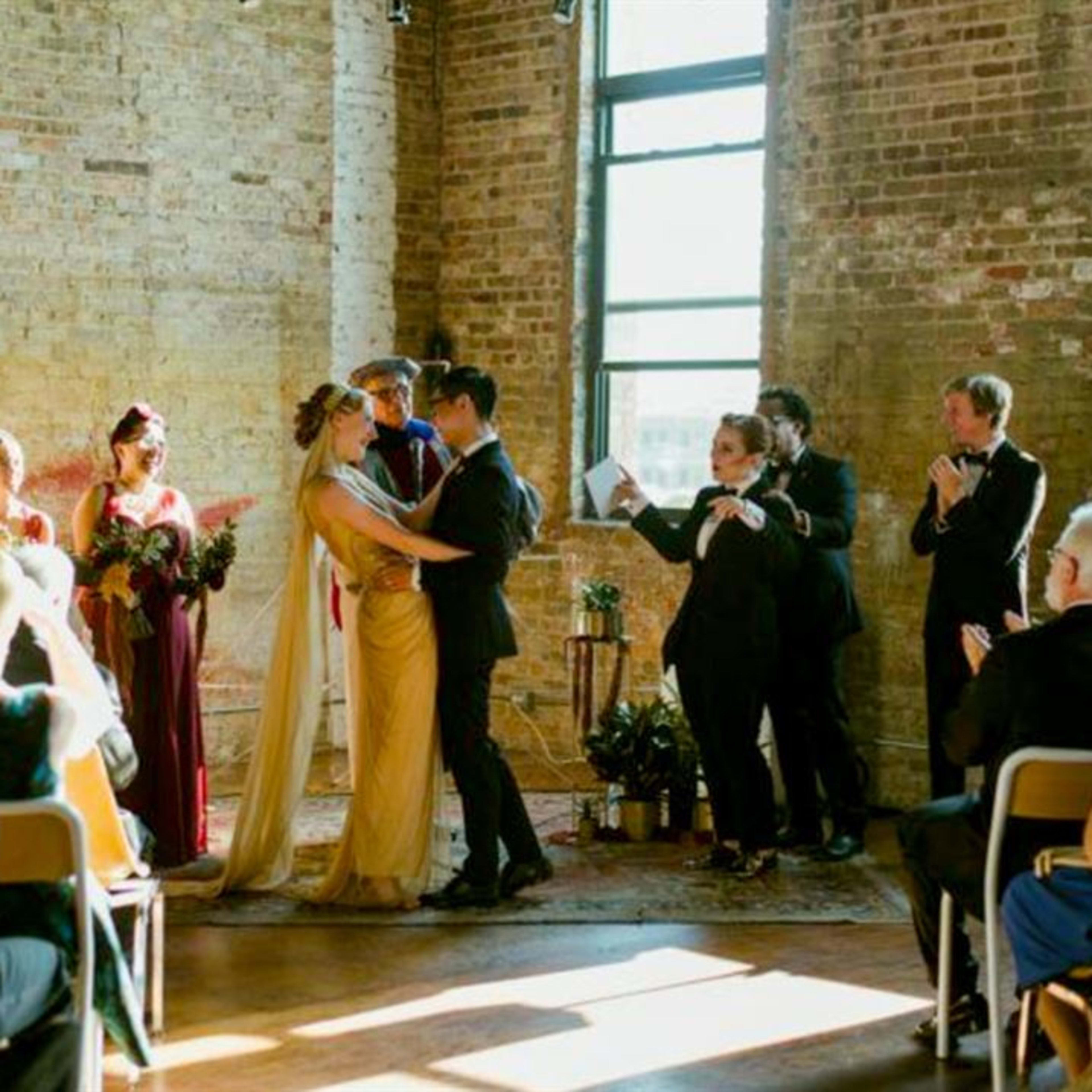 A couple exchanges vows during a wedding ceremony surrounded by their wedding party and guests in a rustic venue.