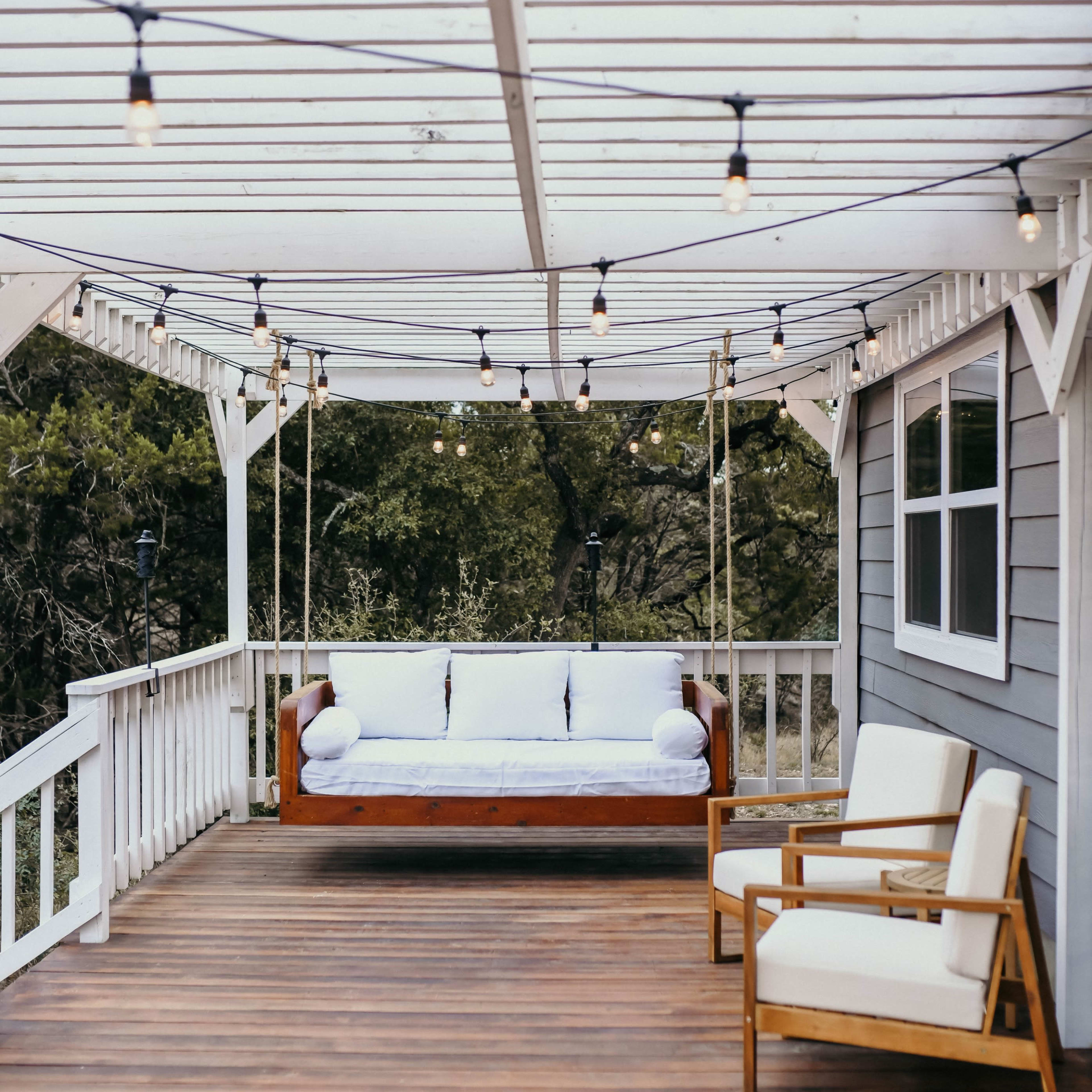 A covered wooden deck with a swing bench and two white cushioned chairs, adorned with string lights overhead.