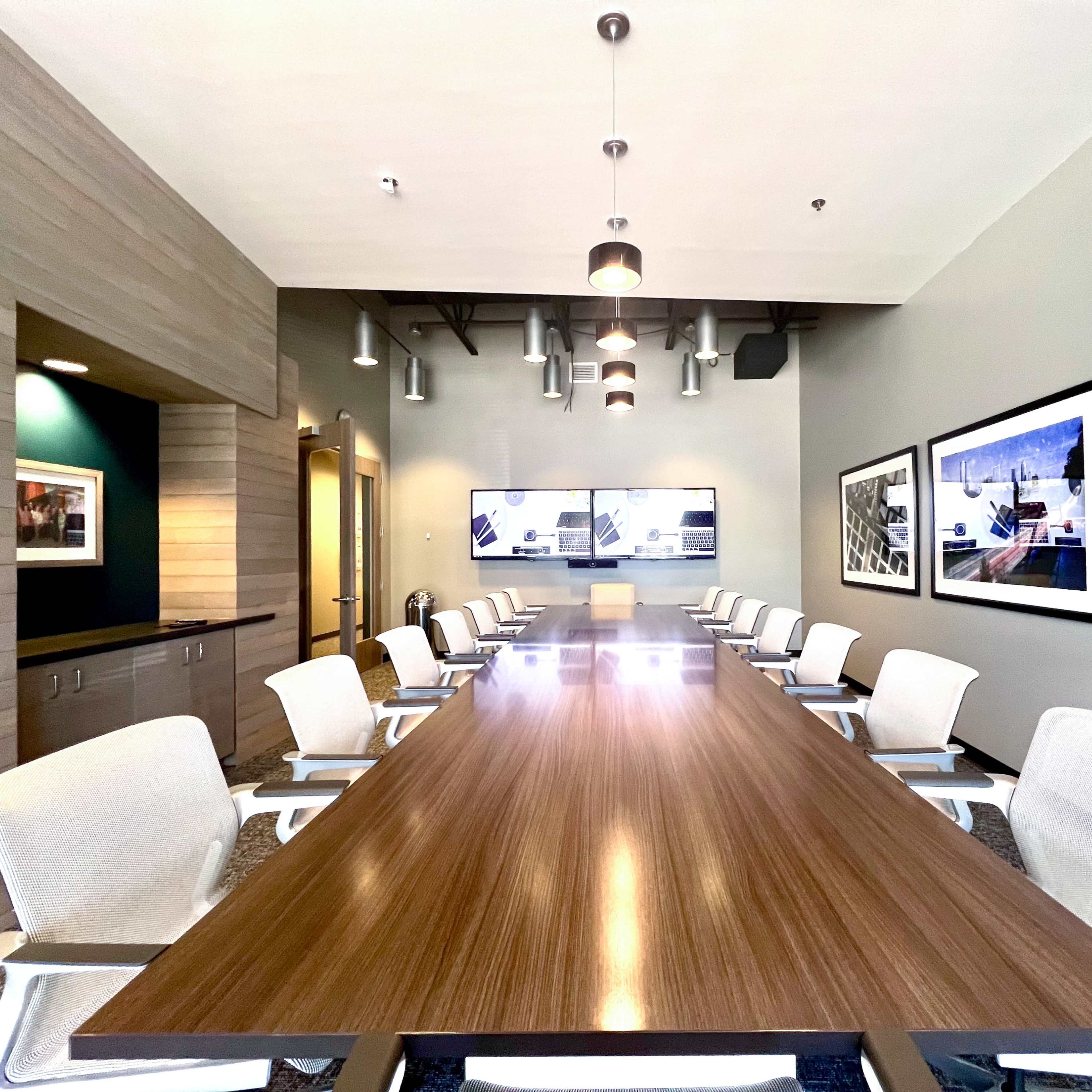 A long conference table with white chairs is set in a modern meeting room featuring wall-mounted screens and framed photographs.