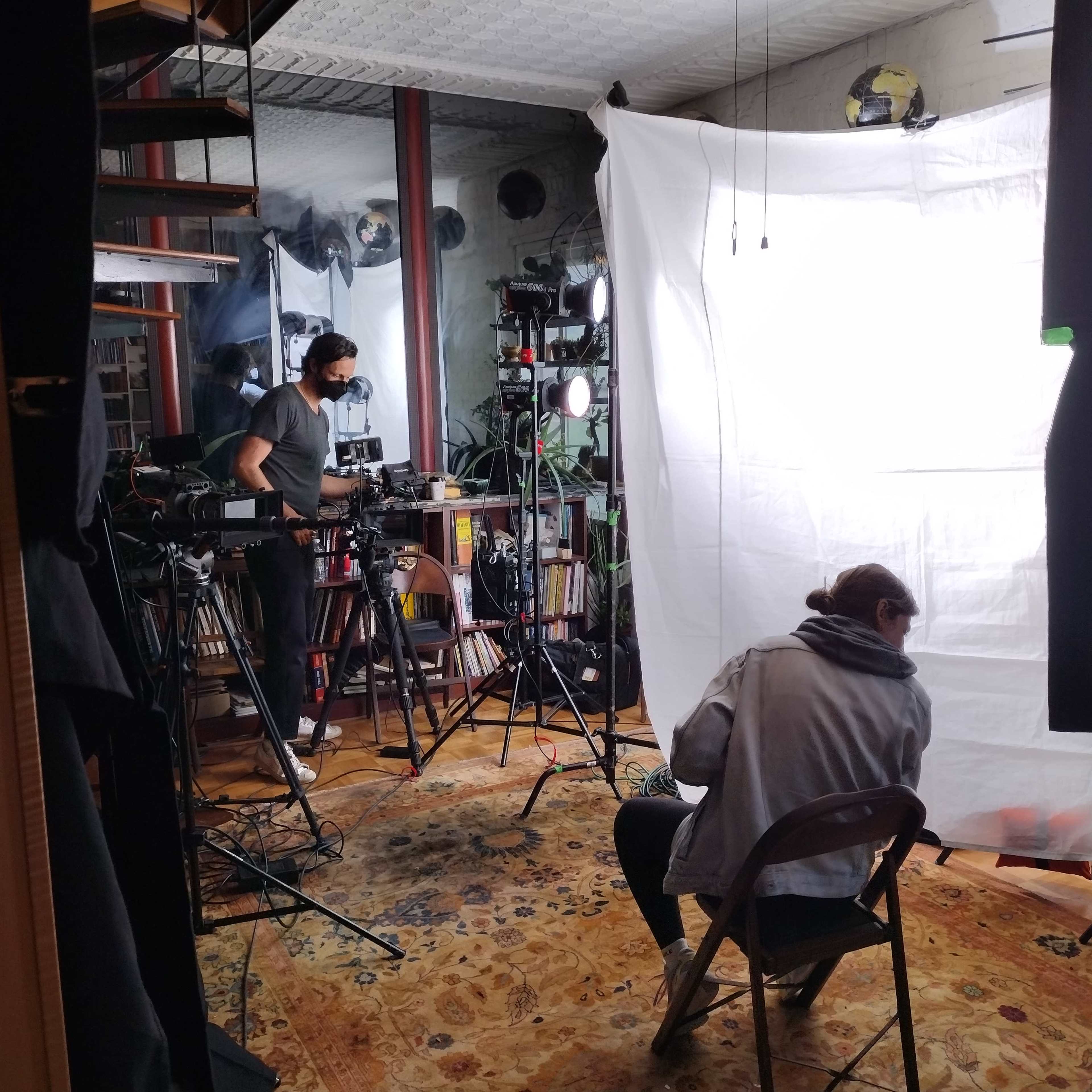 A film setup is shown in a room with two cameras on tripods and a person seated in front of a white backdrop.