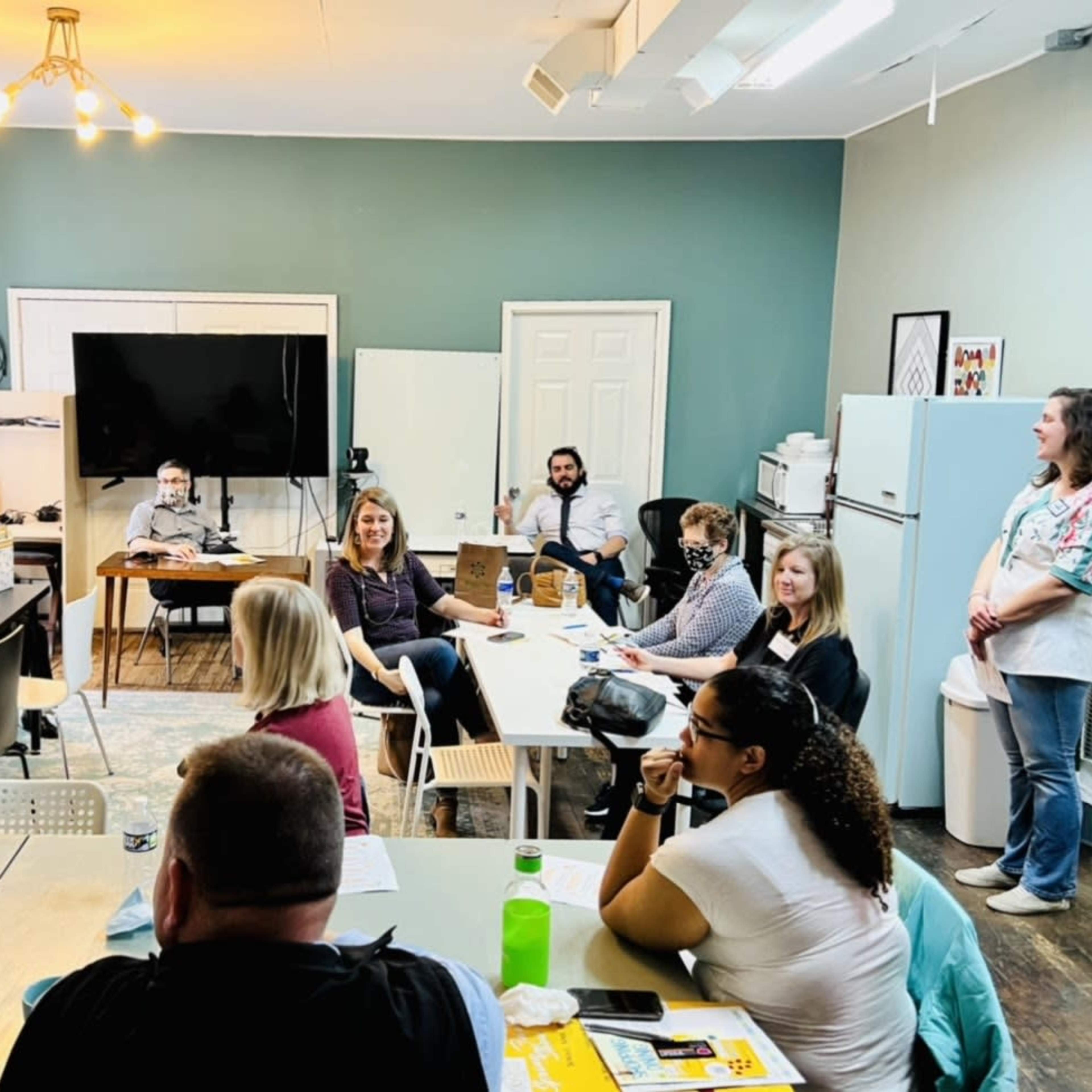 A group of people sit at tables in a well-lit meeting room with a television and whiteboard at the front.