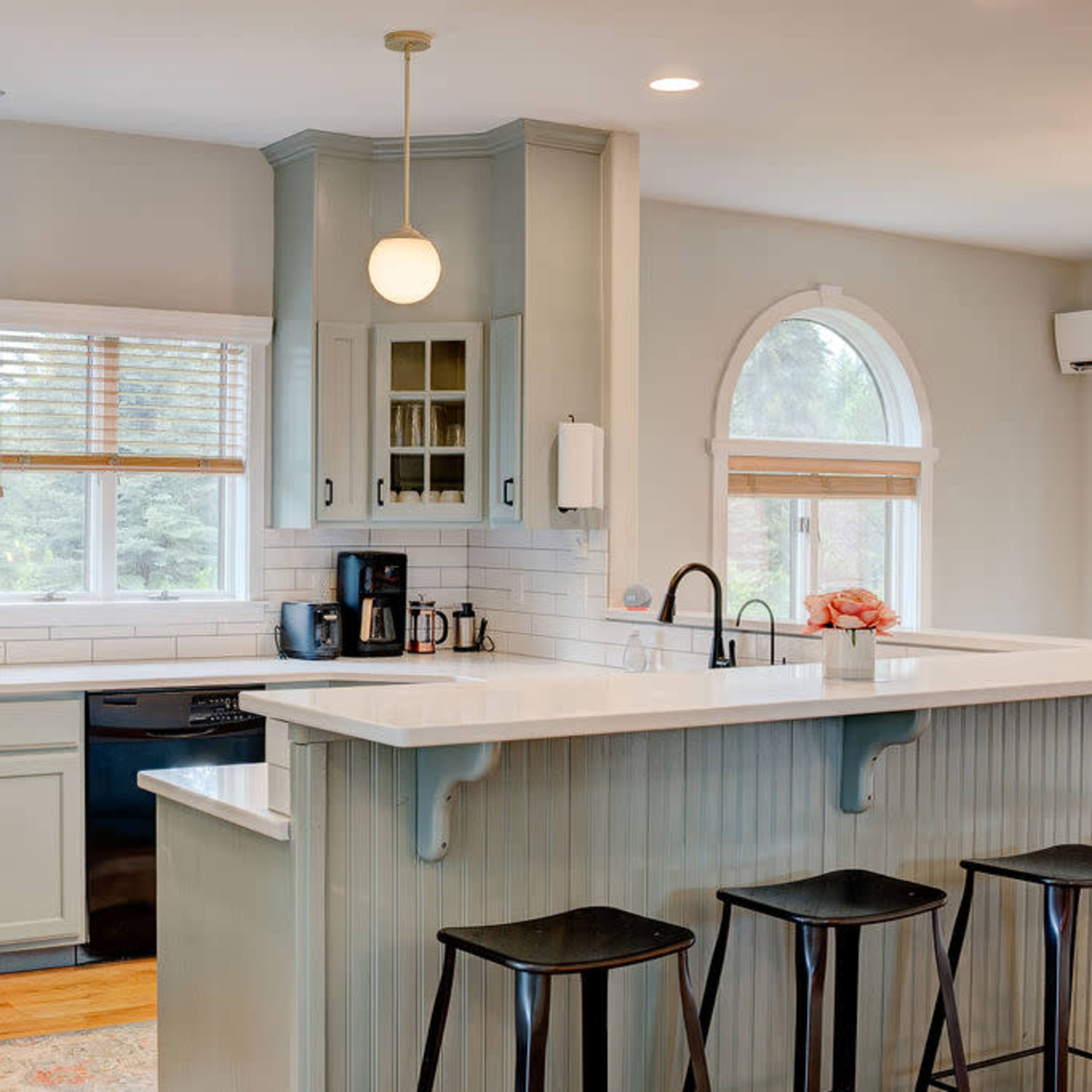 A modern kitchen with light blue cabinetry, a white countertop, and three black bar stools at the island.