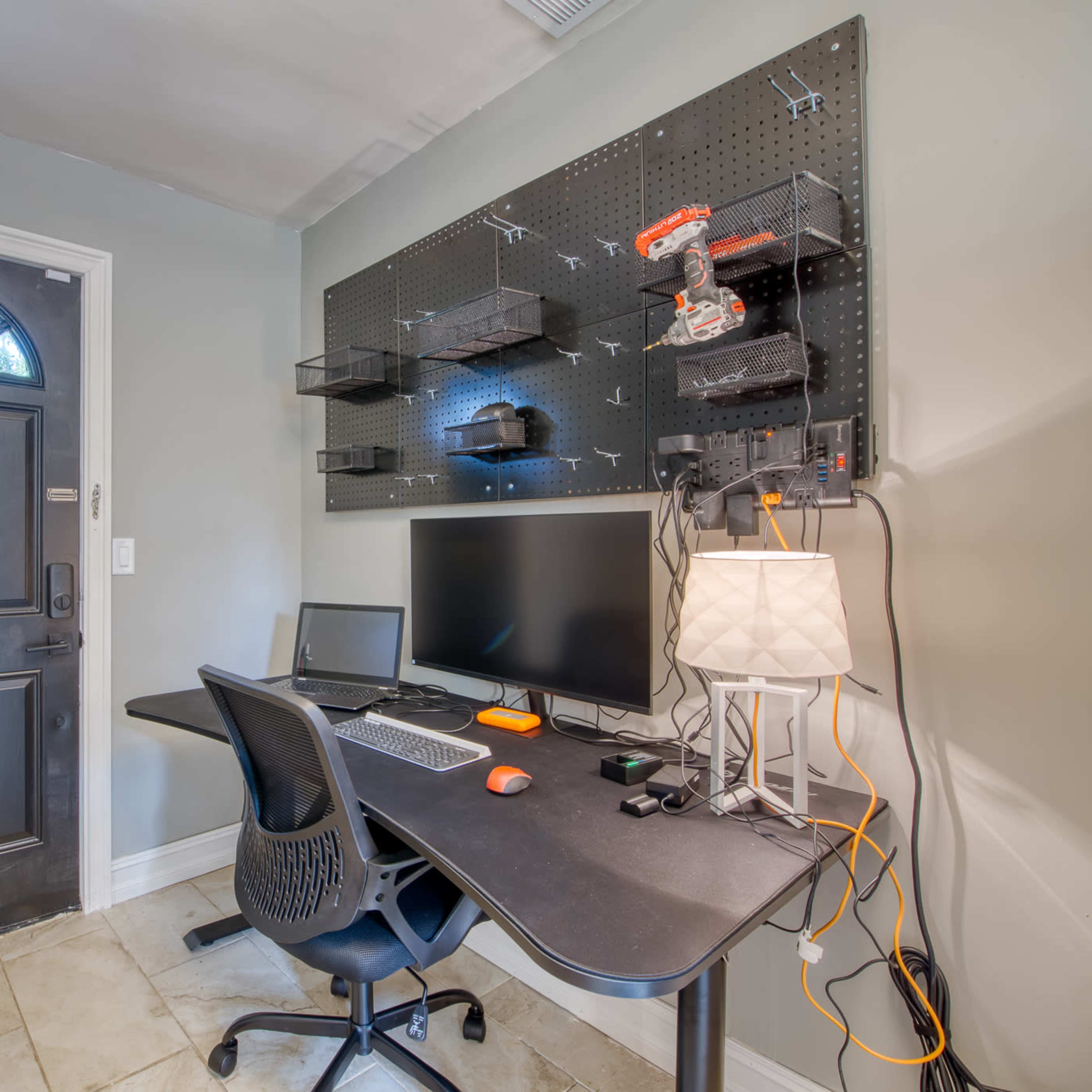 A small office space features a desk with a computer, a lamp, and a pegboard wall with various tools, next to a front door.