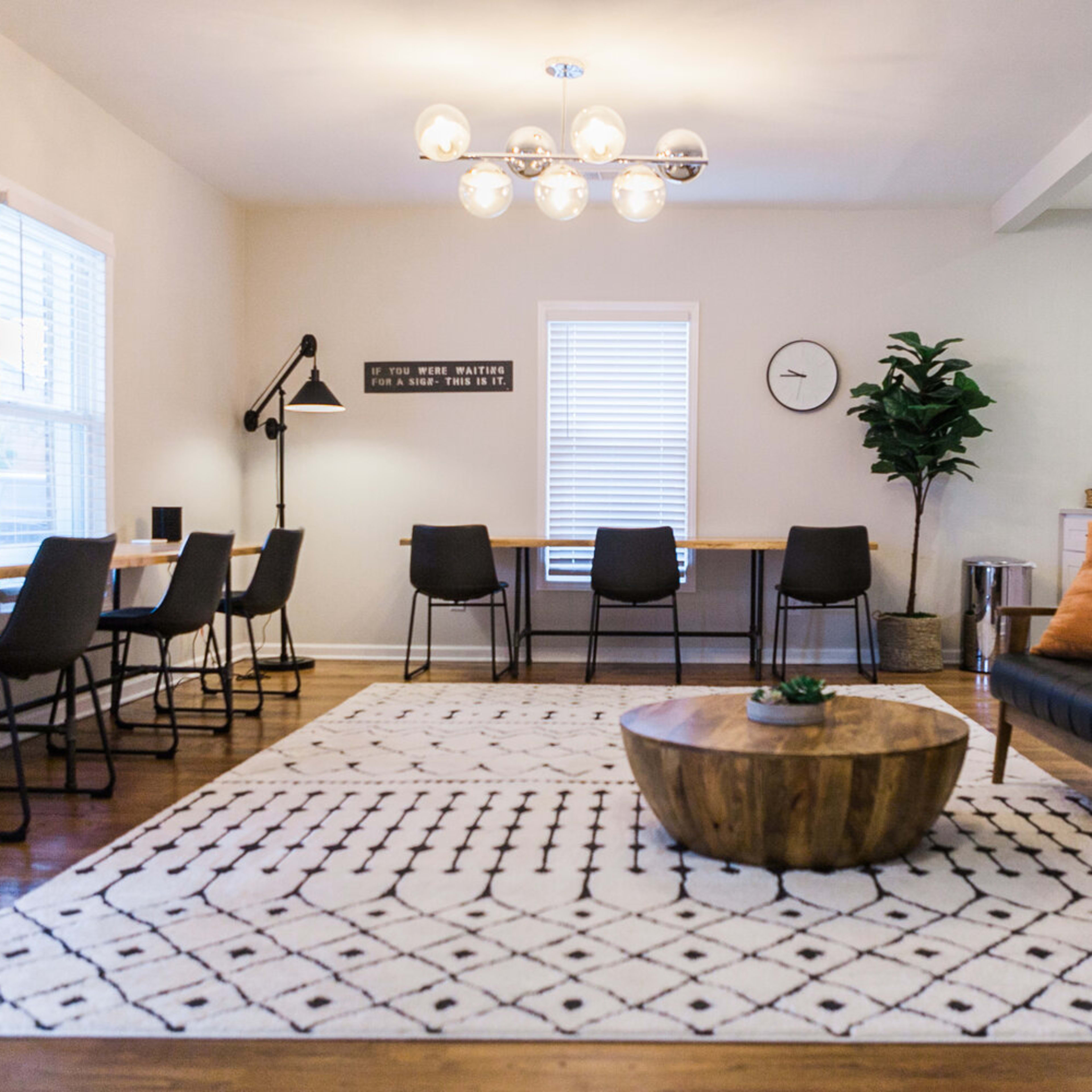 A modern living and dining area featuring a wood coffee table, a patterned area rug, several black chairs, and a large light fixture overhead.