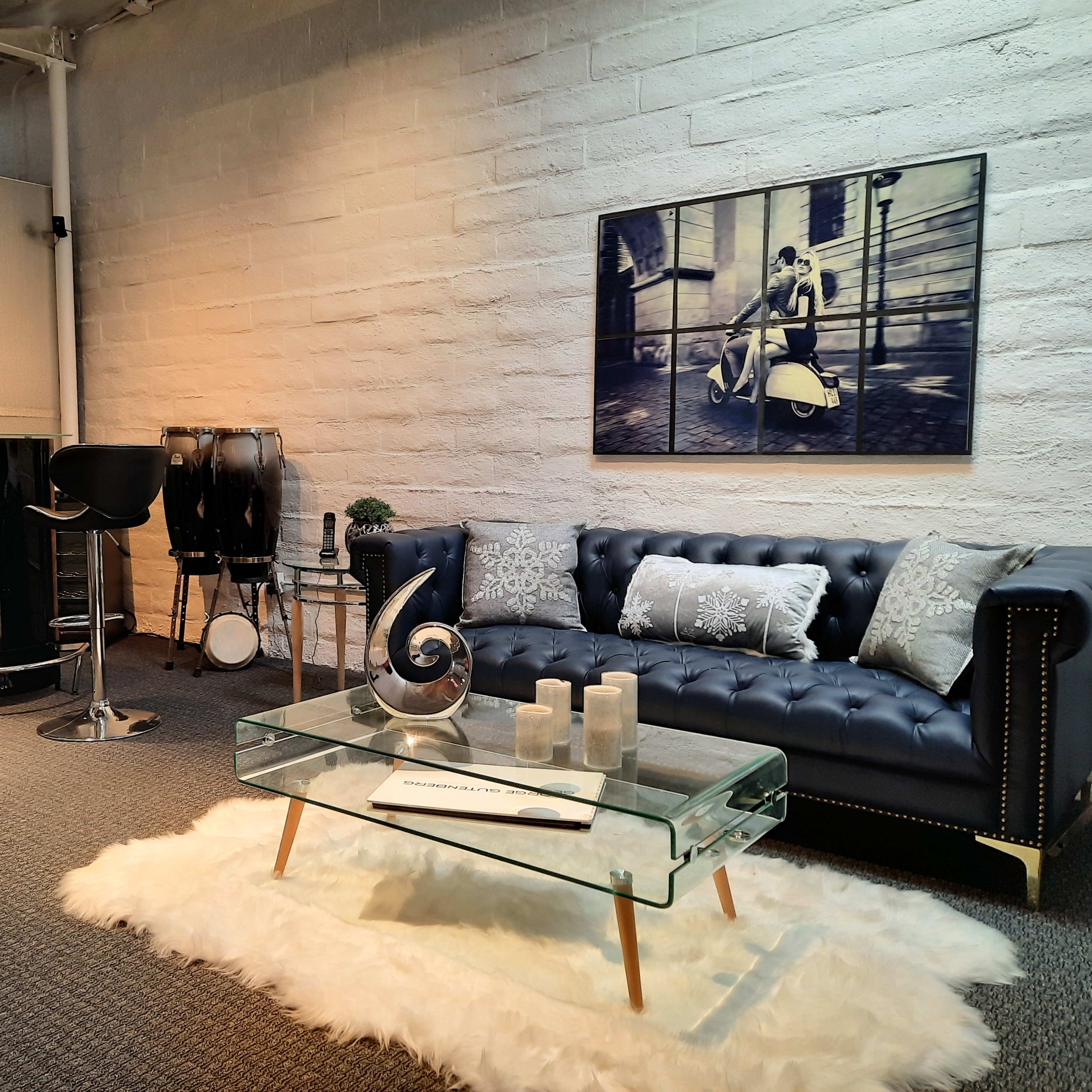A modern lounge area featuring a dark sofa, a glass coffee table on a white rug, and bar stools arranged around a counter.