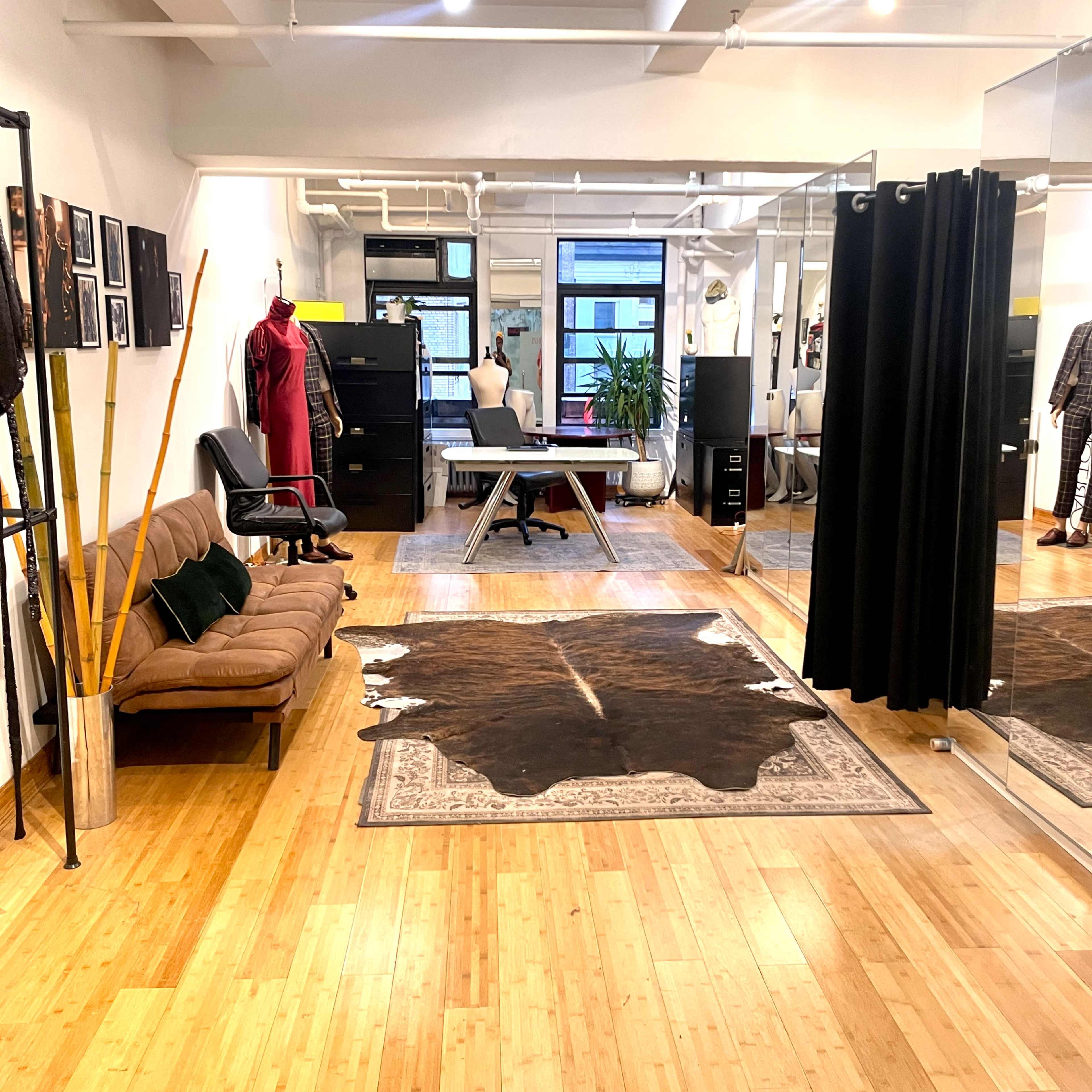 A spacious studio with wooden floors, a brown sofa, a large mirror, a black curtain, a tailored work table, and a decorative cowhide rug.
