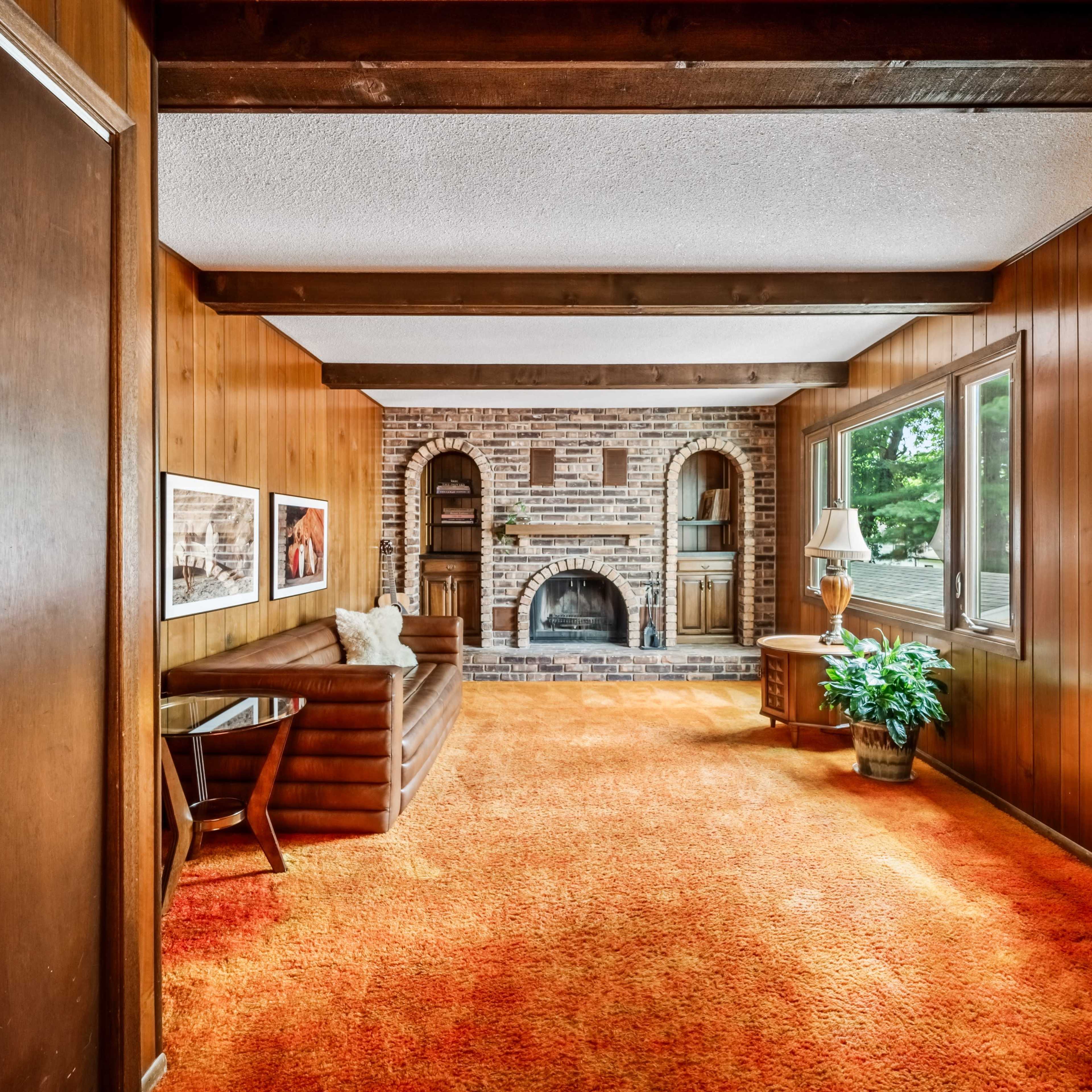 A living room with wooden paneling, a brick fireplace, a couch, and large windows letting in natural light.
