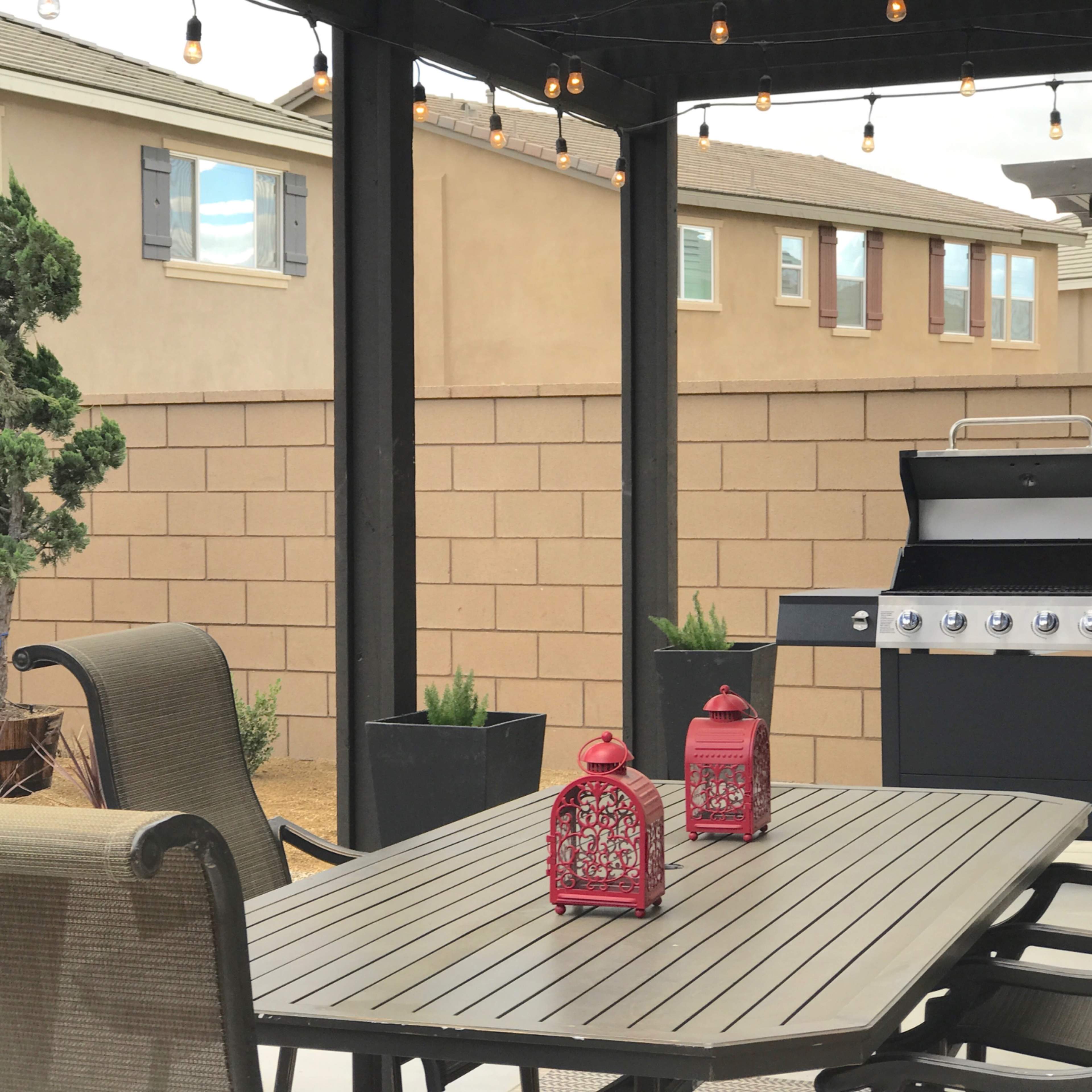 A patio setup features a table, chairs, red lanterns, and a grill under a pergola, with a fenced backyard in the background.