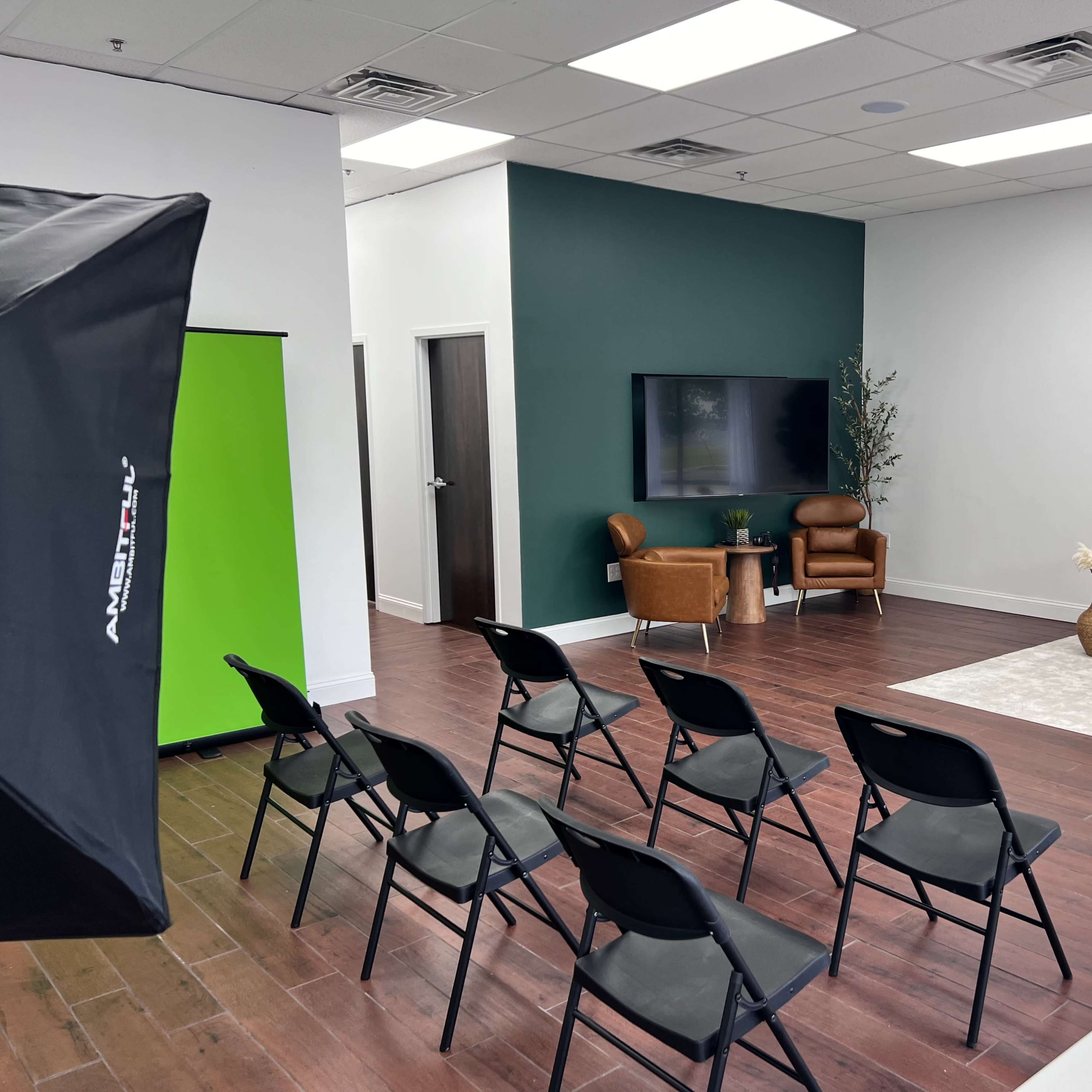 A modern, minimalistic interior with a seating arrangement of black chairs facing a light-colored sofa and a television against a green accent wall.