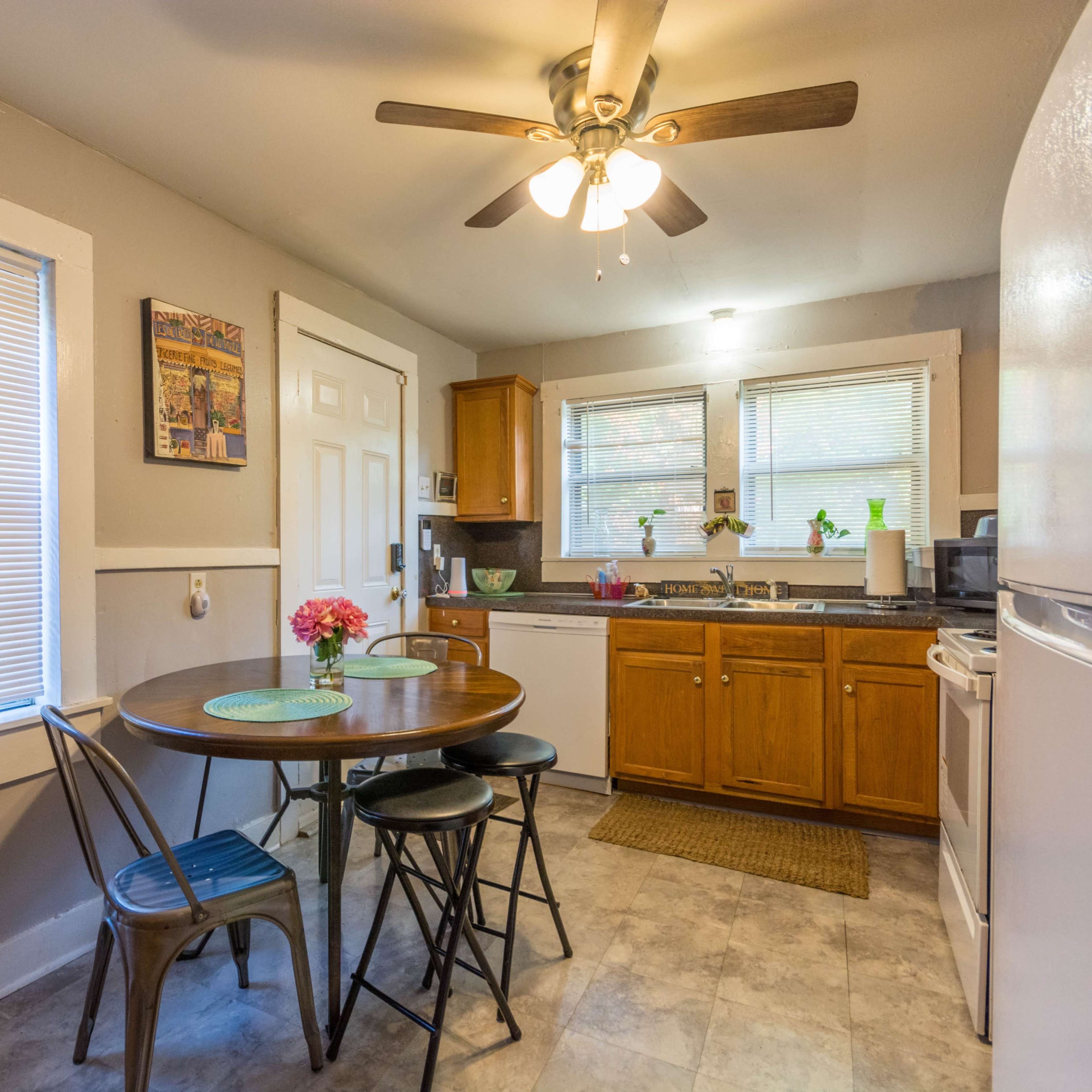 A small kitchen features wooden cabinets, a round dining table with two chairs, a ceiling fan, and white appliances.