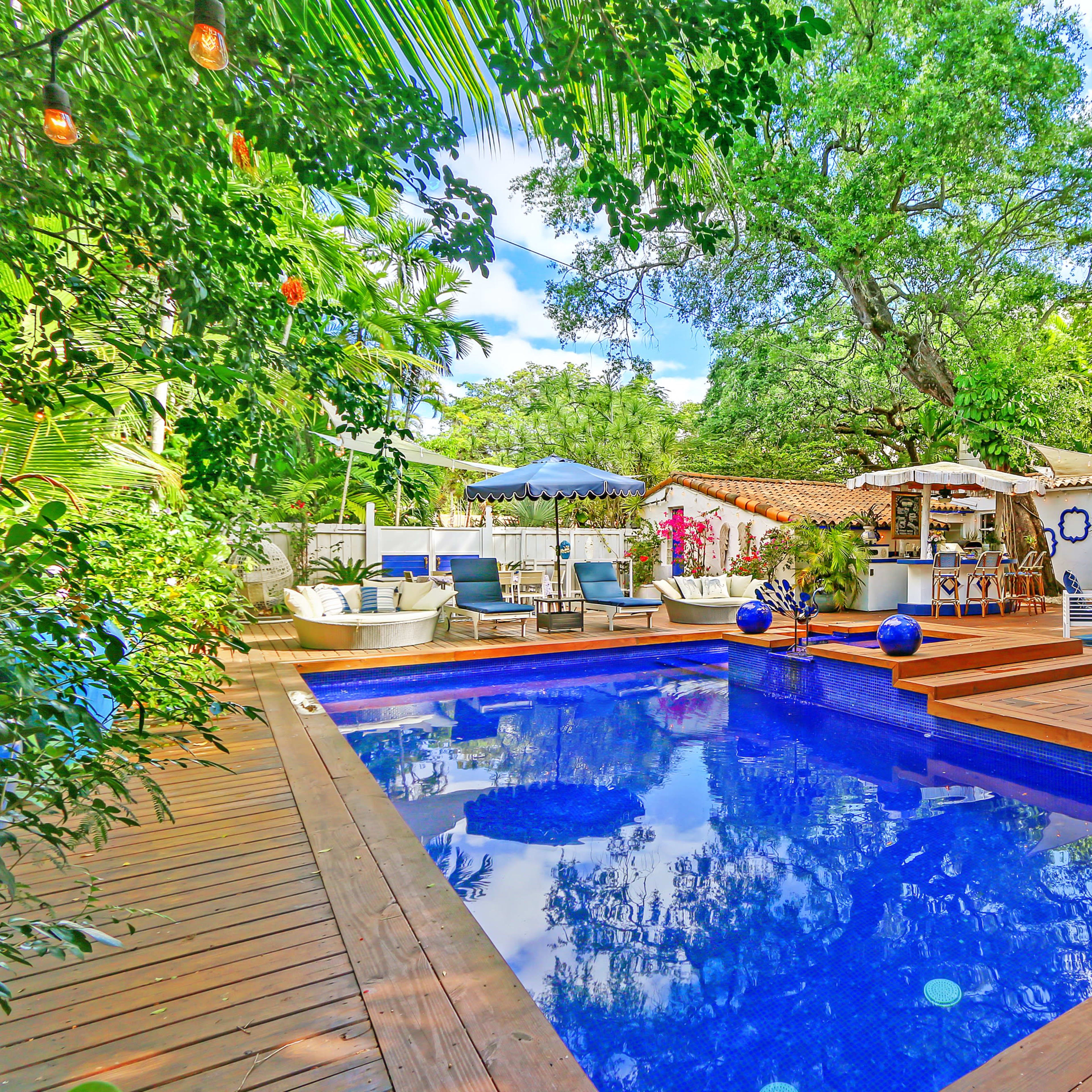 A rectangular swimming pool surrounded by wooden decking, lounge chairs, and lush greenery, with a patio area and various outdoor furnishings nearby.