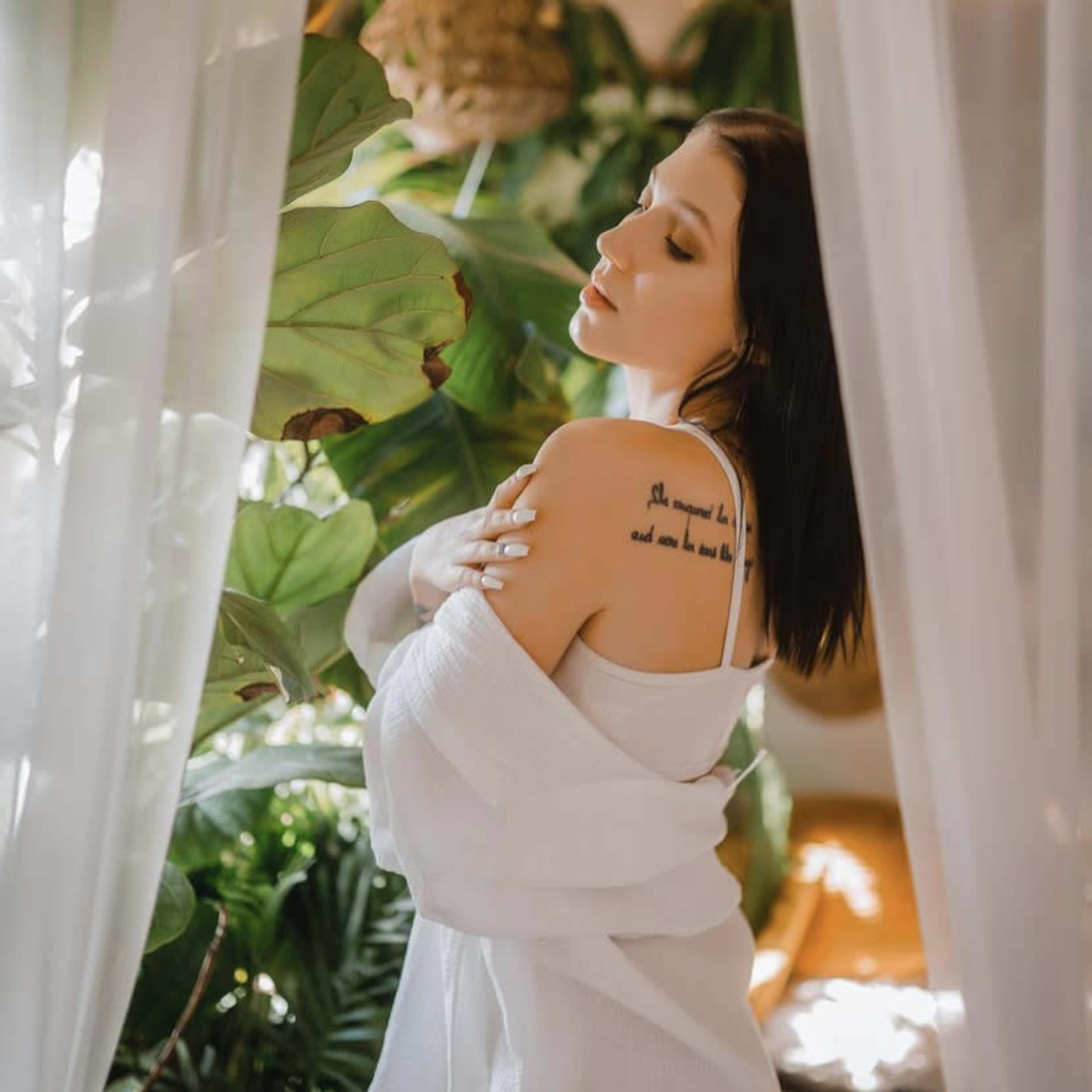 A woman in a white outfit poses beside lush green plants, with sheer curtains framing the scene.