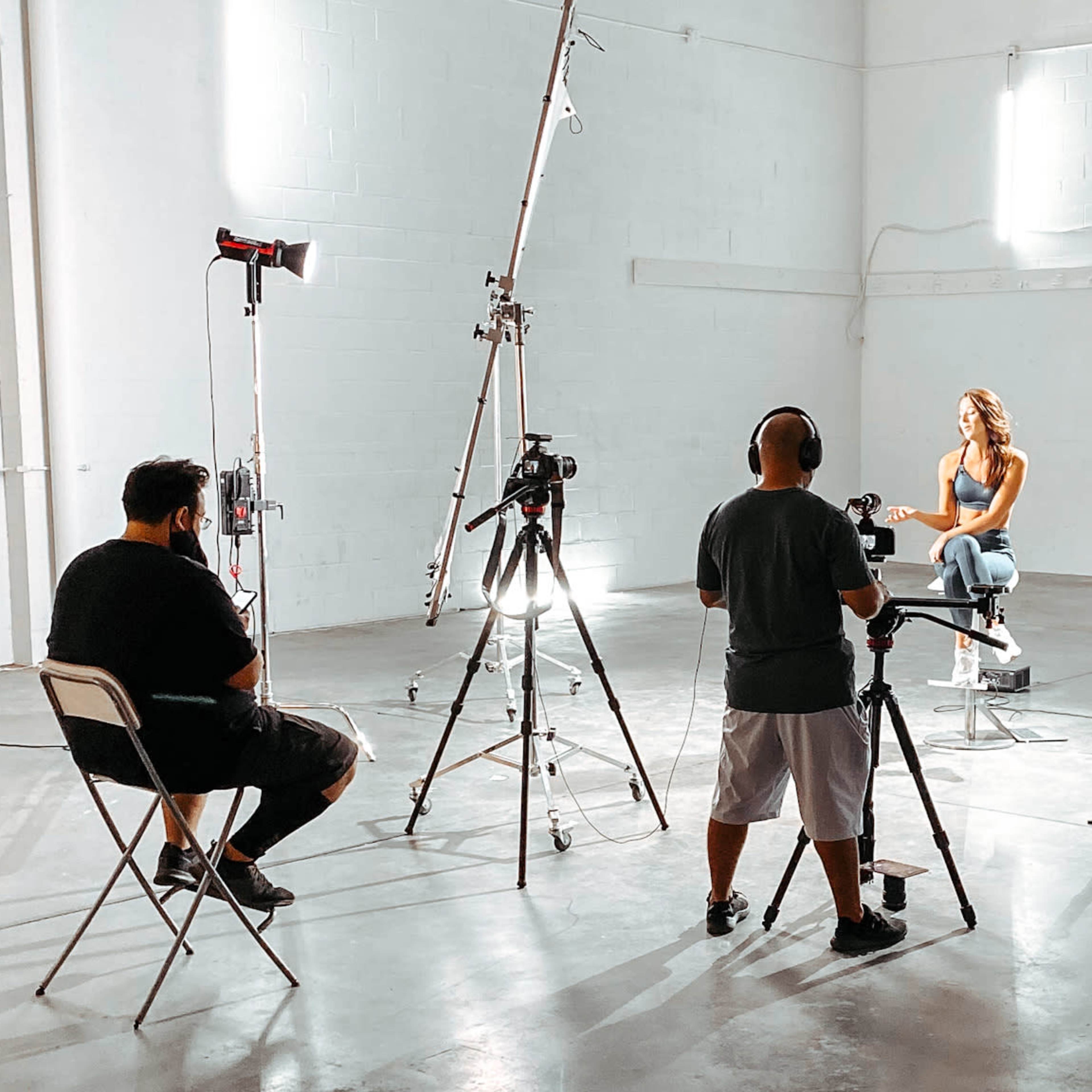 A woman sits on a stool in a brightly lit studio, while three individuals operate cameras and lighting equipment around her.