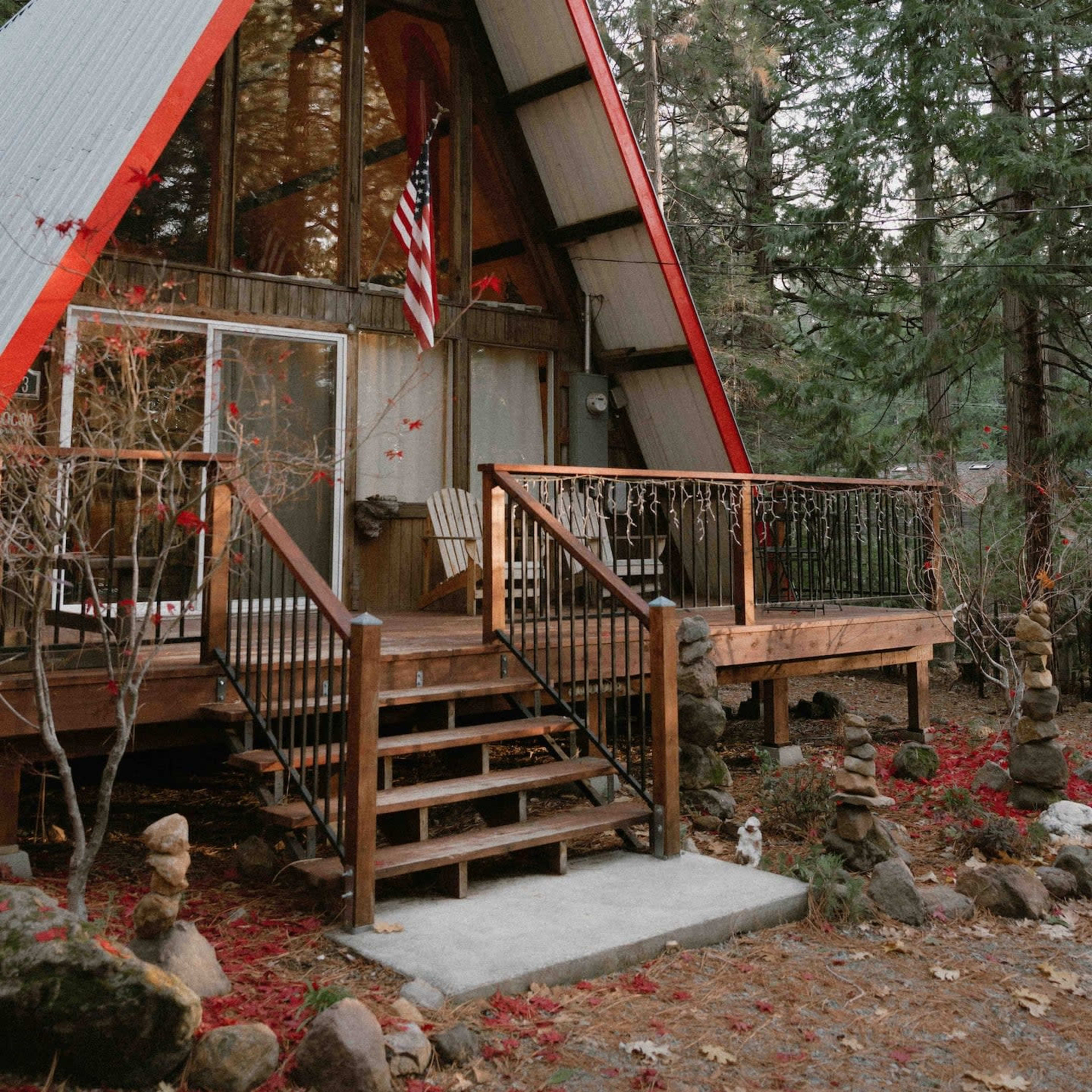 An A-frame cabin with a red roof, nestled among tall trees and featuring a wooden deck with steps and an American flag.