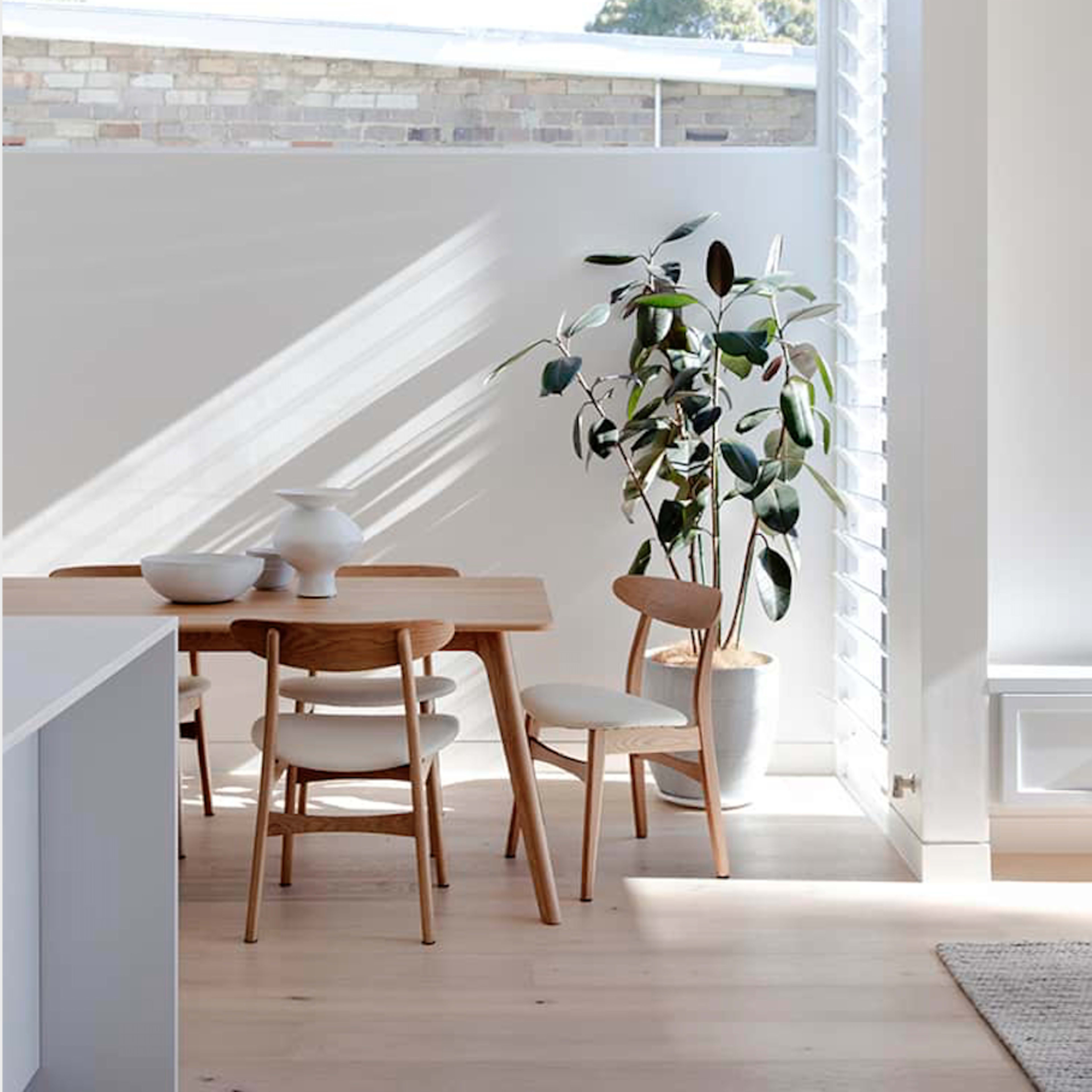A wooden dining table with three chairs and a potted plant is positioned near a window that lets in natural light.
