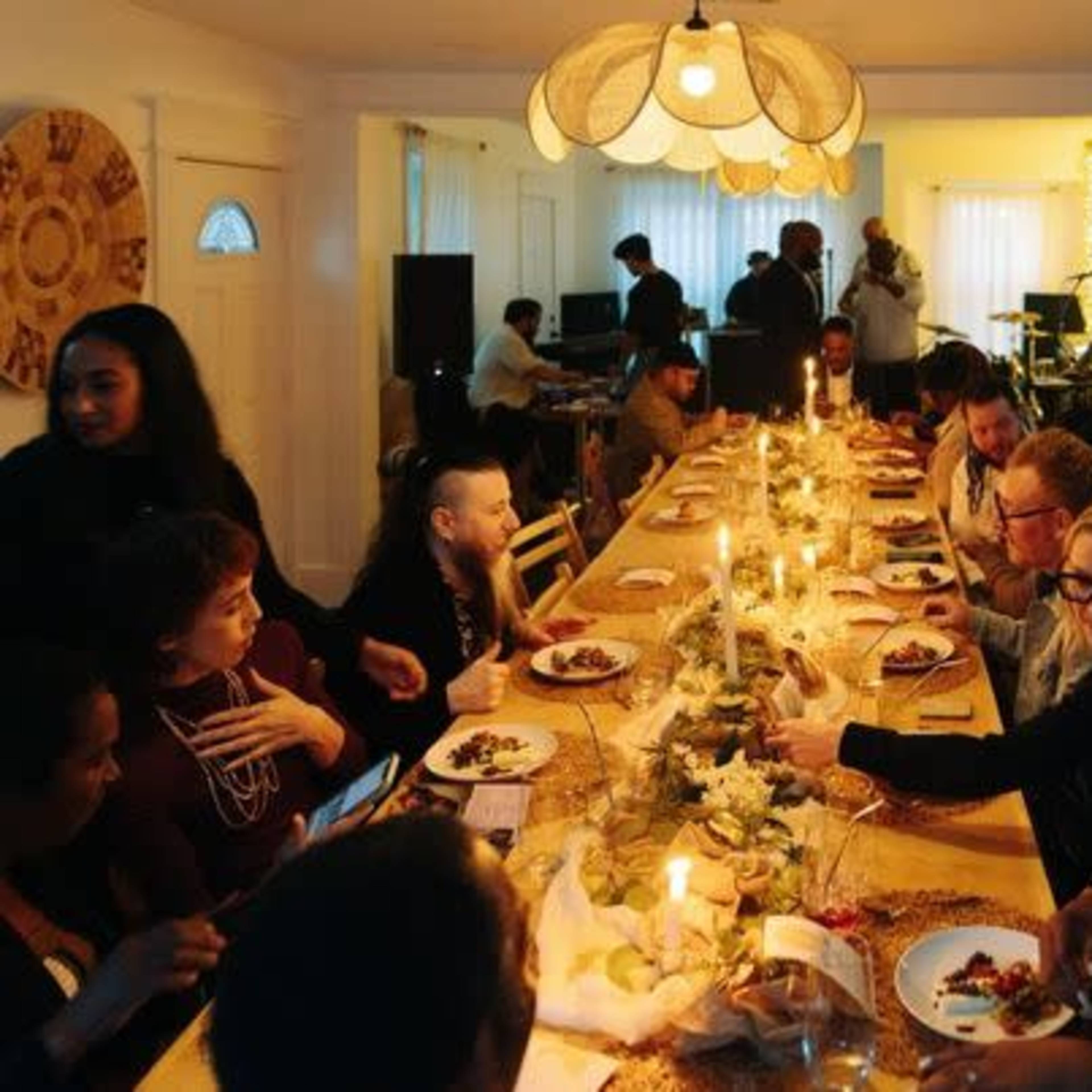 A long dining table is set with plates of food and candles, while a group of people enjoy a meal and converse in a brightly lit room.