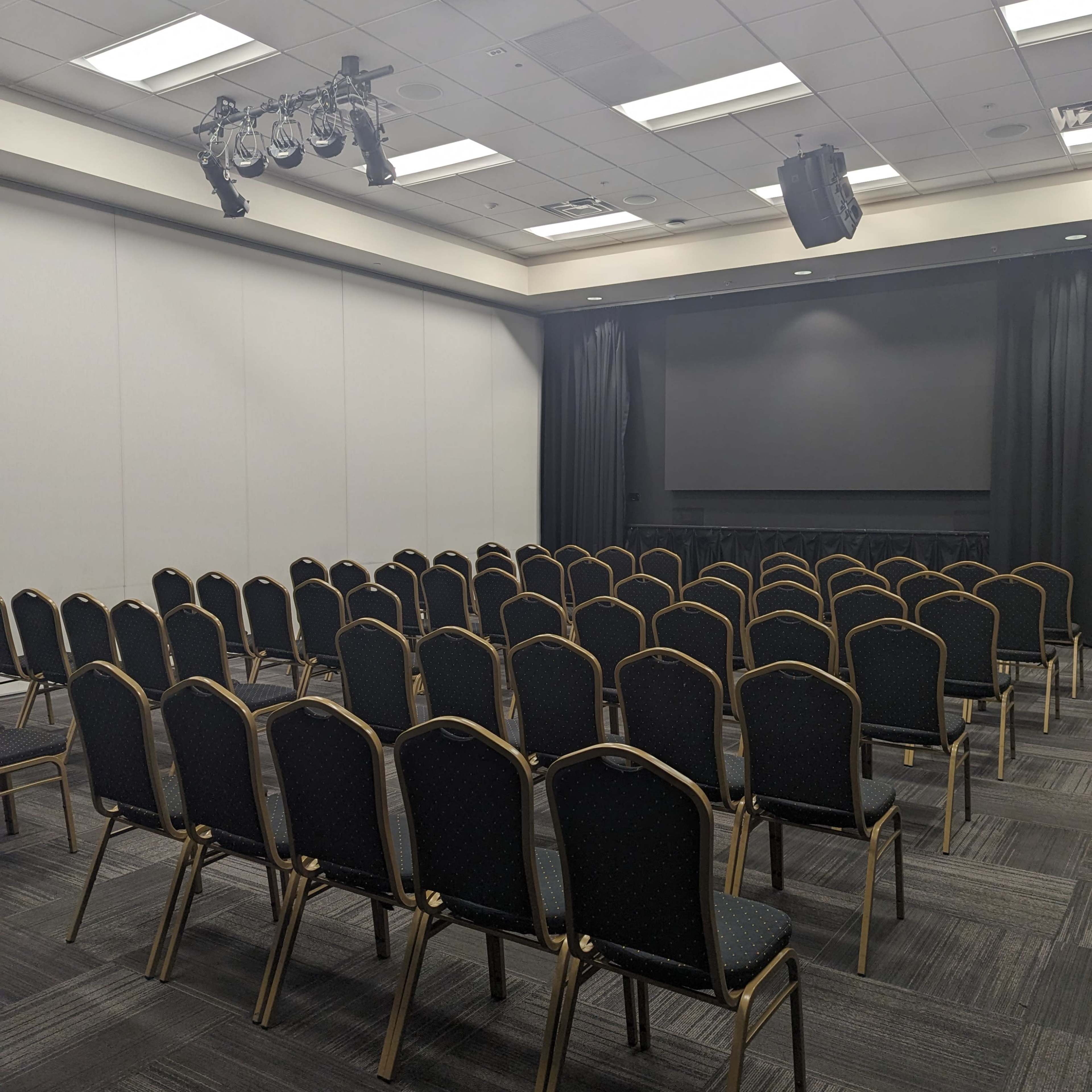 A conference room set up with rows of empty black chairs facing a stage area with curtains and lighting.